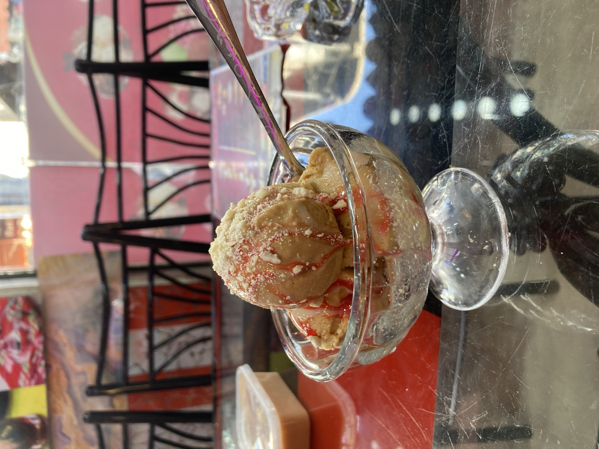 A close-up shot depicts a glass dessert bowl filled with light brown ice cream or gelato, topped with red sauce and crushed crumbs. A silver spoon is partially submerged in the ice cream. The bowl rests on a reflective surface, potentially a table. In the background, partially visible, are red and black patterned chairs and other glass containers. The photo was taken indoors, possibly in a restaurant or cafe. It is plausible that the image was taken in Quetta City Tehsil, Pakistan.