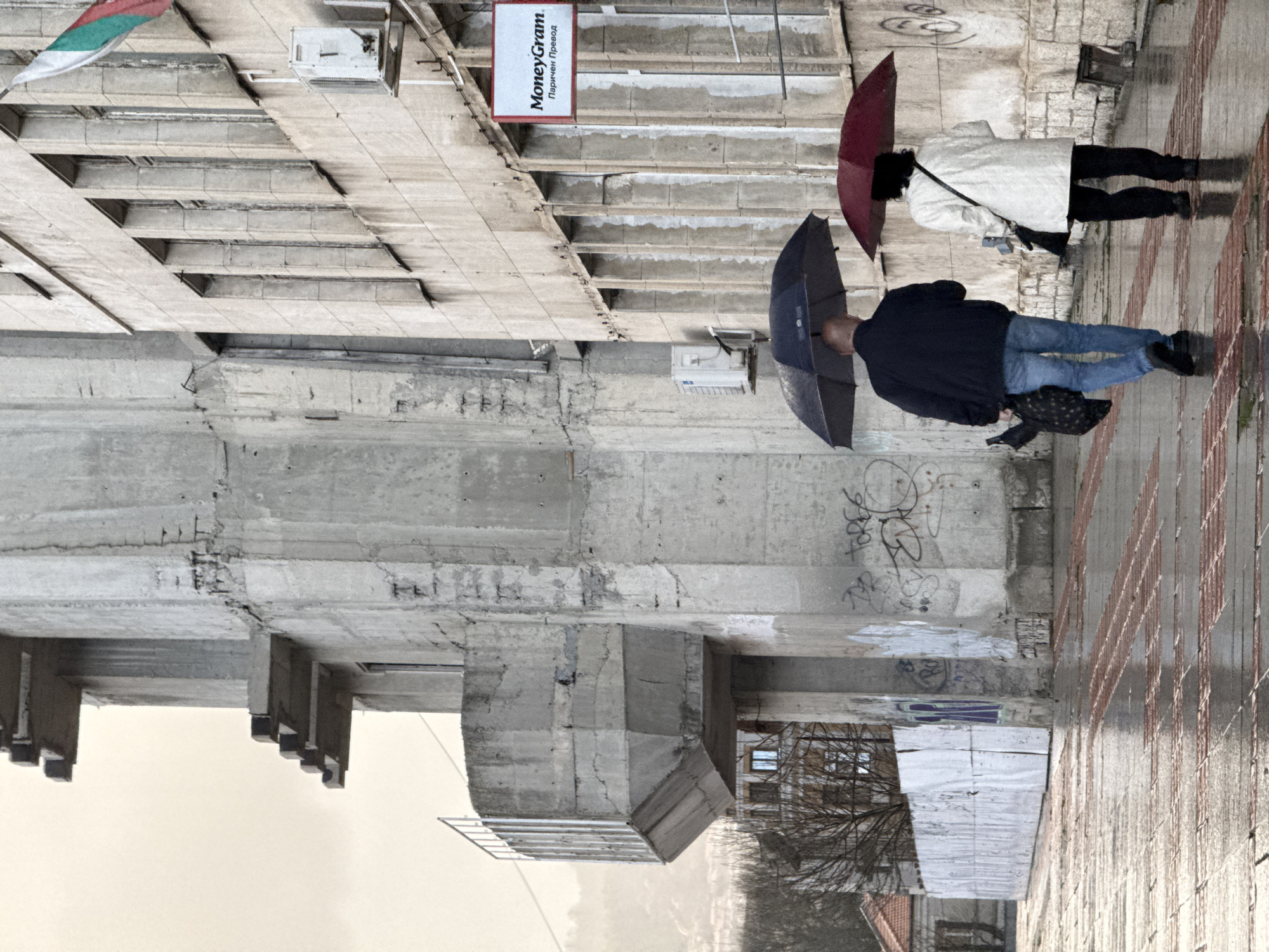 The image shows a street scene in Shumen, Bulgaria on a rainy day. The perspective is rotated 180 degrees, so the sky appears at the bottom. Two people walk with umbrellas: one person in a black jacket and blue jeans holding a dark umbrella, and another in a light-colored coat and black pants holding a red umbrella. The sidewalk is wet and reflects the scene. A modern concrete building is adjacent to the sidewalk. A MoneyGram sign is visible above a doorway. A Bulgarian flag hangs from the building. Graffiti marks a section of the concrete structure. The street is paved with tiles. Distant trees and buildings are visible.