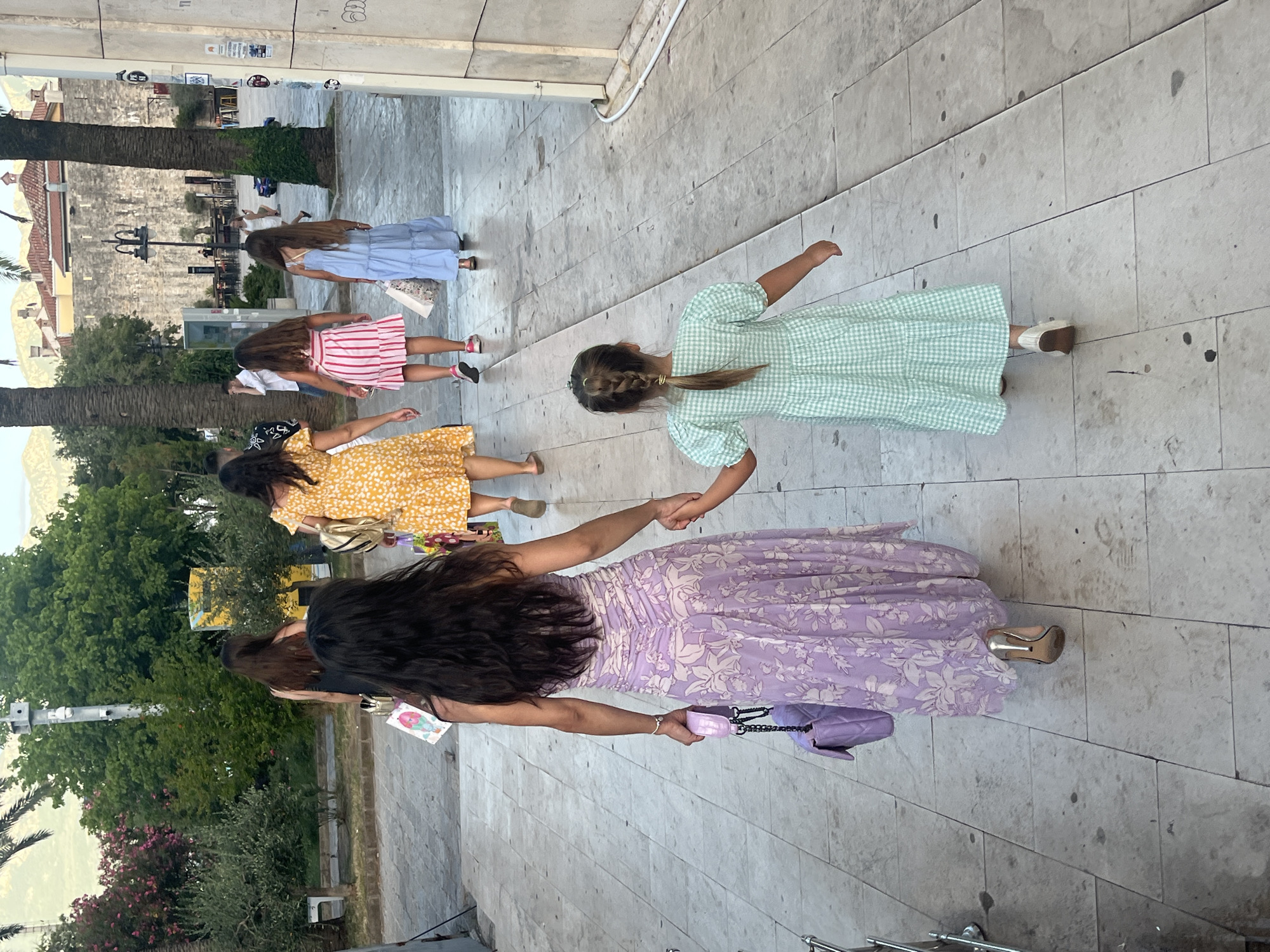 In Budva, Montenegro, a group of individuals walks along a stone-paved pedestrian walkway lined with palm trees. A woman in a long, floral purple dress and high heels walks hand-in-hand with a young girl wearing a green checkered dress. Behind them, other women and girls walk ahead. In the background, a stone wall and buildings are visible.