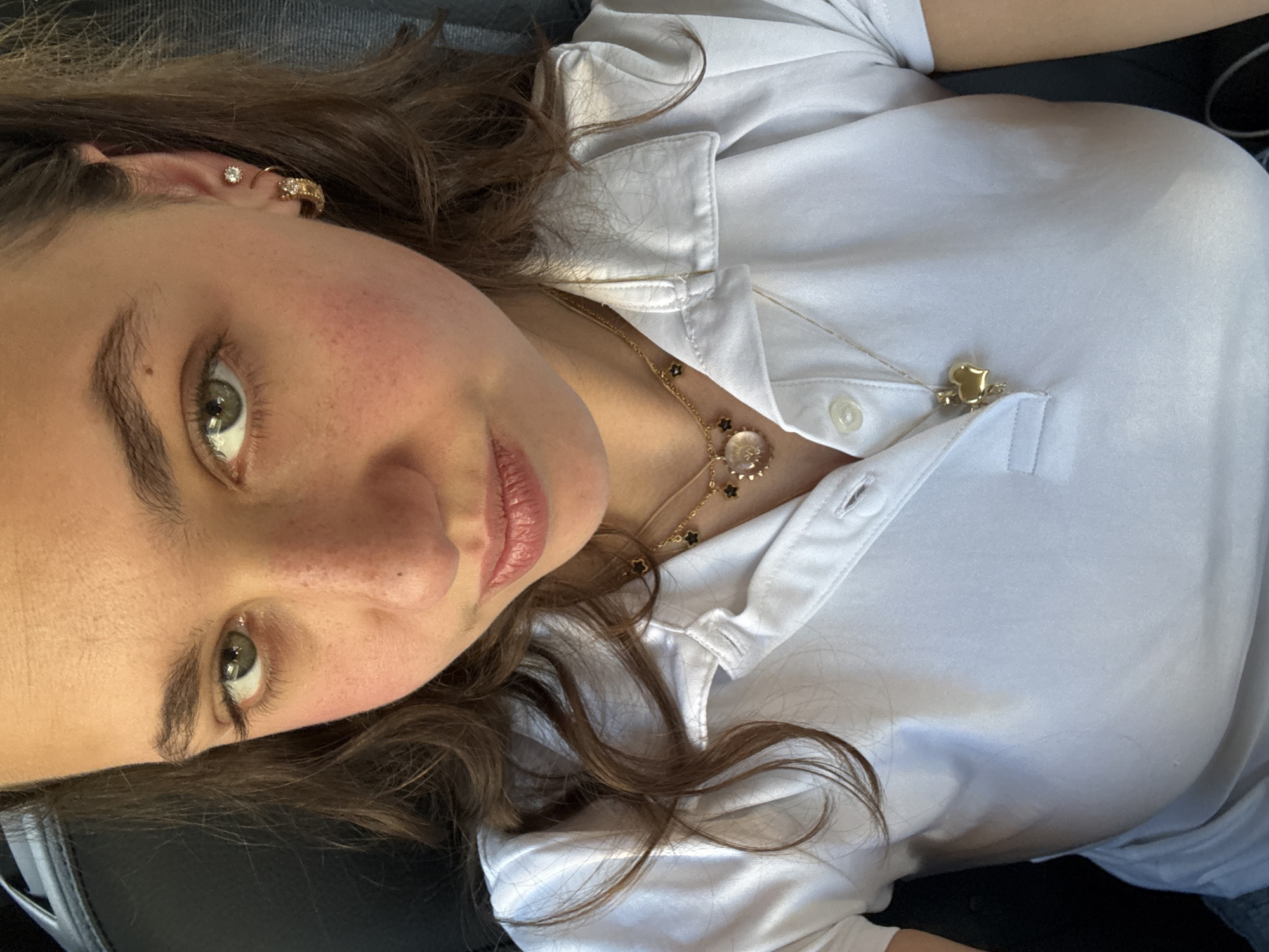 A close-up, tilted shot shows a person with fair skin, green eyes, and brown hair wearing a white collared shirt. The person has freckles and multiple earrings in their left ear. Two gold necklaces are visible, one with flower-shaped pendants and the other with a heart-shaped pendant on the shirt's collar. The person is likely indoors, seated in a dark interior. The location within Milan, Italy, cannot be determined from the image.