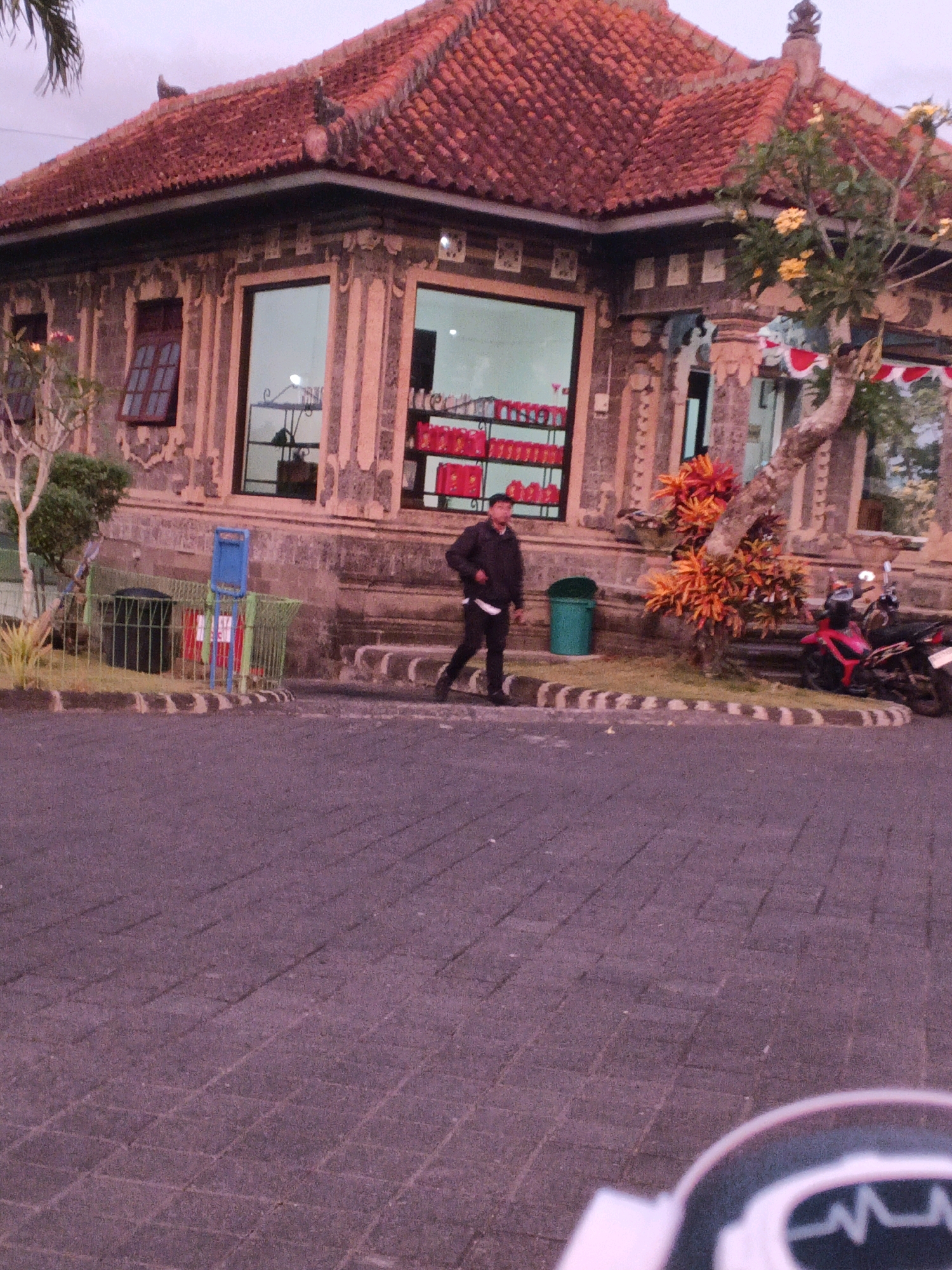 The image depicts a street scene in Denpasar, Indonesia, with a man walking in the foreground on a paved walkway. Behind him is a building with a red tiled roof and ornate stone facade, featuring large windows. A motorcycle is parked near a tree with colorful foliage. The overall atmosphere suggests an outdoor commercial or residential area.