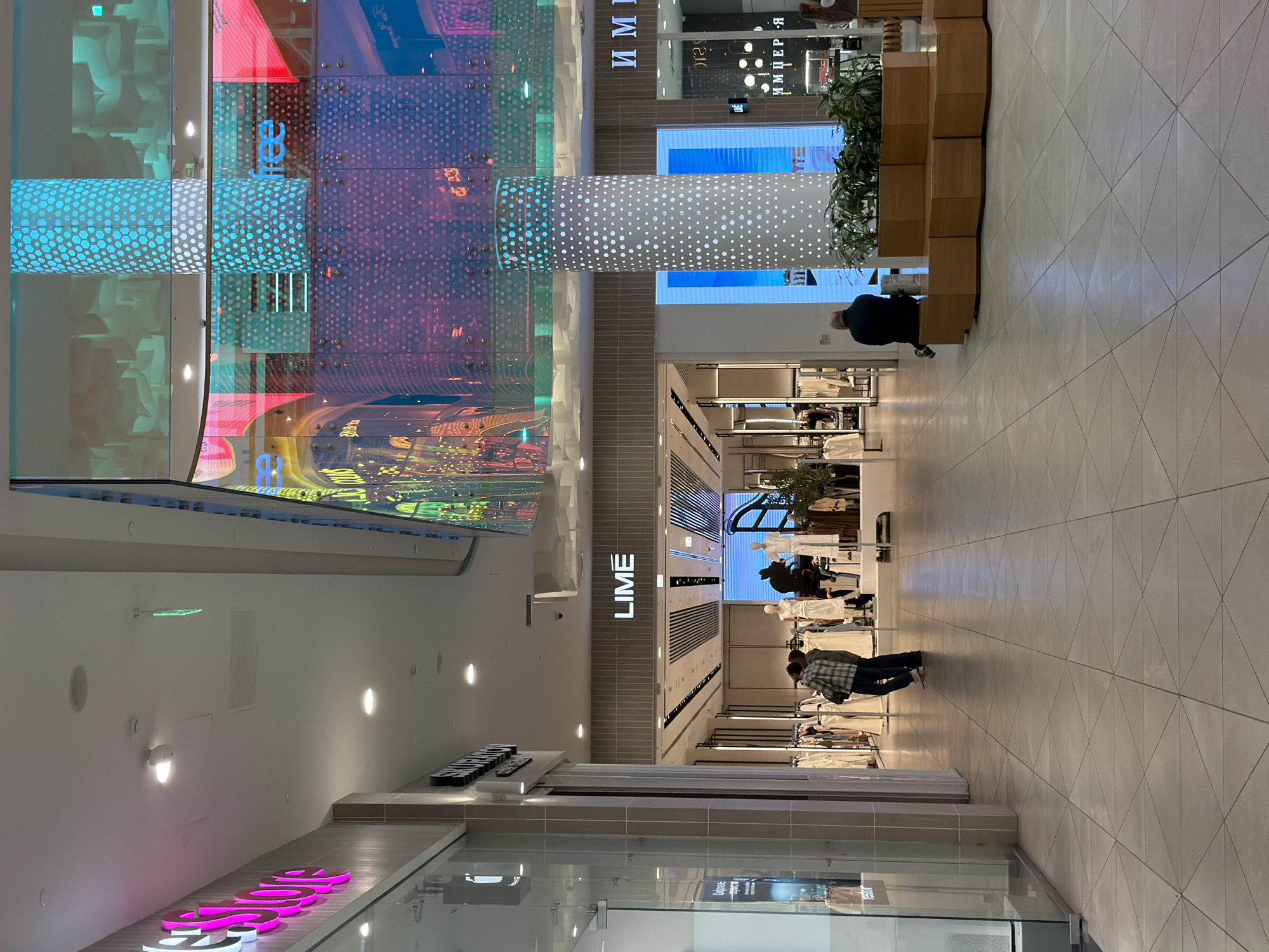 The image shows an interior view of a shopping mall in Kazan, Russia. The mall has a high ceiling with a curved, illuminated panel featuring a colorful, abstract design. A large cylindrical pillar with dotted patterns supports the ceiling. The floor is tiled in a light color with a geometric pattern.
Retail stores are visible on both sides of the hallway. Signs for "LIME" and "ИМПЕРИЯ" are present. Several shoppers are browsing through the clothes on display racks within the store. A person is sitting on a wooden bench with plants in the background.