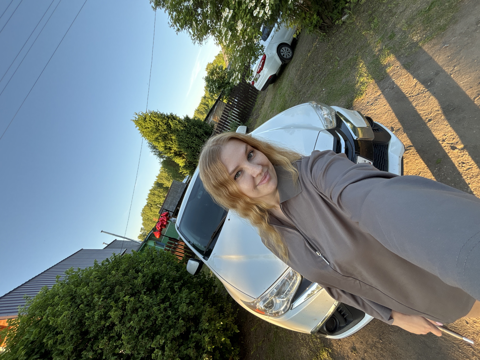 The image depicts a blonde female taking a selfie in a rural or semi-rural residential setting within the city of Rozhdestveno, Russia, under a clear blue sky, likely during late afternoon or early evening based on the angled sunlight and long shadows. The woman, with mid-length wavy blonde hair and light eyes, is wearing a long-sleeved grey-brown top with a zipper and is smiling directly at the camera. She is positioned in front of a white Mitsubishi ASX crossover, which is angled slightly left, with its front bumper, grille, and headlights prominently visible.
In the background to the left of the woman, dense green foliage obscures part of a corrugated metal building. Above the crossover's front, a cluster of red heart-shaped balloons is visible near a wooden picket fence and a smaller, older-looking building. To the right of the woman, a white sedan (possibly a Kia Rio) is parked further back on an unpaved dirt path, with its rear and side visible. A bush with white blossoms stands between the woman and the sedan. Numerous green trees form a dense treeline in the background. The ground is an unpaved dirt or gravel surface with patches of grass and weeds. Utility lines traverse the upper right portion of the clear blue sky.