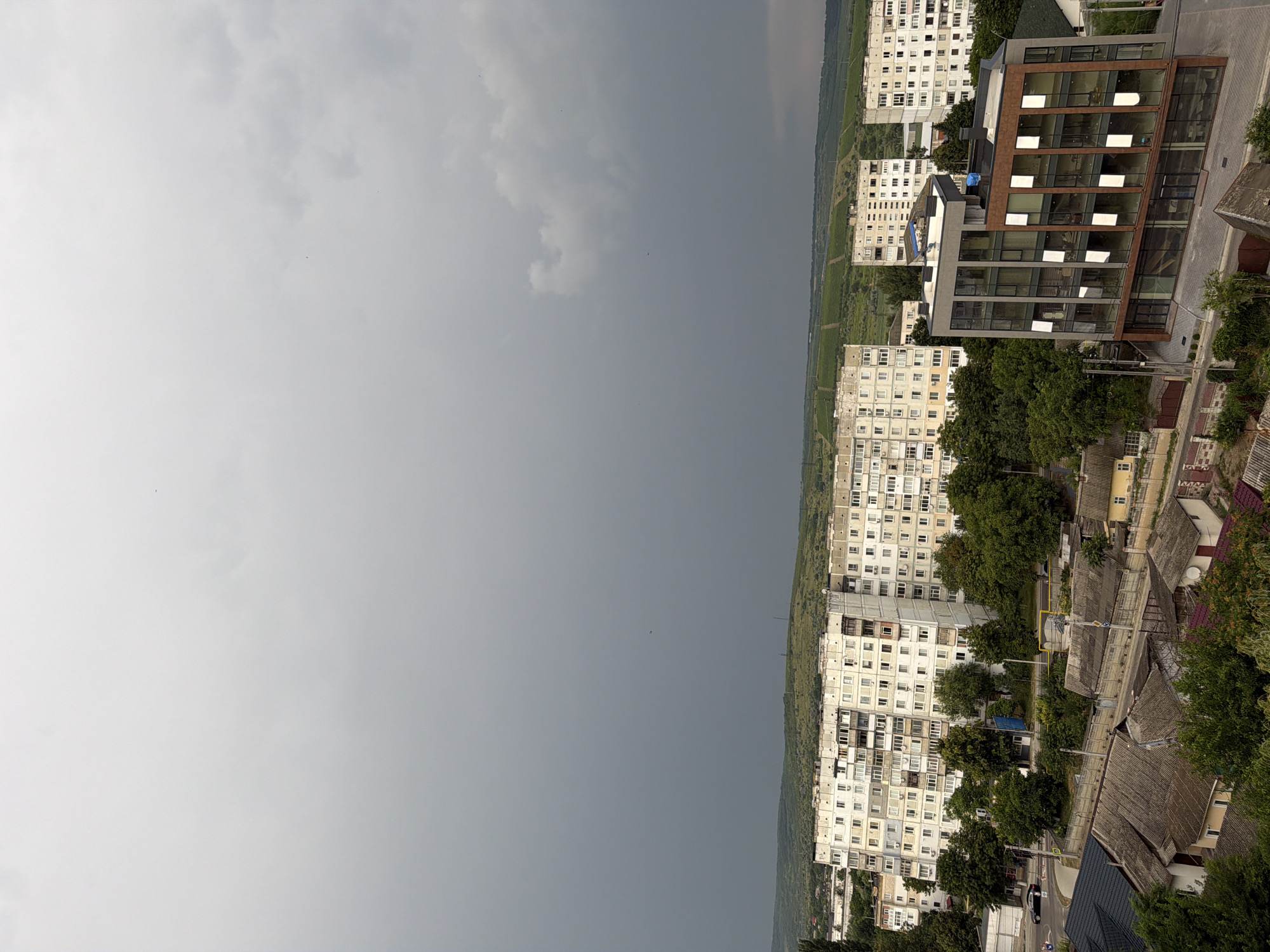 This is a medium-angle shot of a cityscape in Strășeni, Moldova, under a heavily overcast sky. The foreground features a cluster of older, low-rise houses with grey, pitched roofs. To the left, a black car is parked on a paved road, with a glimpse of a woman walking on a sidewalk. Behind these houses, several large, multi-story apartment buildings, characteristic of Soviet-era architecture, dominate the middle ground. These buildings are light-colored with numerous balconies and windows. A modern, multi-story commercial building with a glass facade is positioned to the right of the apartment buildings. In the background, rolling green hills with terraced vineyards are visible, with the dark, ominous clouds of an approaching storm hanging low over the horizon. The overall atmosphere is somber due to the weather conditions.