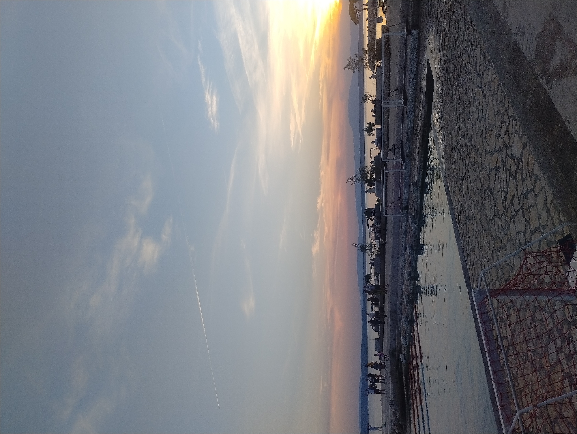 The image depicts a seaside scene in Crikvenica, Croatia, at sunset. A body of water, likely the Adriatic Sea, occupies the lower left portion of the frame, reflecting the sky. The foreground features a paved area constructed from irregular stone slabs, with a white and orange net enclosure, possibly part of a sports field, partially visible in the bottom left corner. Beyond the water, a shoreline is lined with people standing and sitting. A distant, hazy mountain range forms the horizon line. The sky displays a gradient of colors from soft blues and whites at the top to oranges and yellows near the setting sun on the right. A faint contrail from an aircraft is visible in the upper left quadrant of the sky.