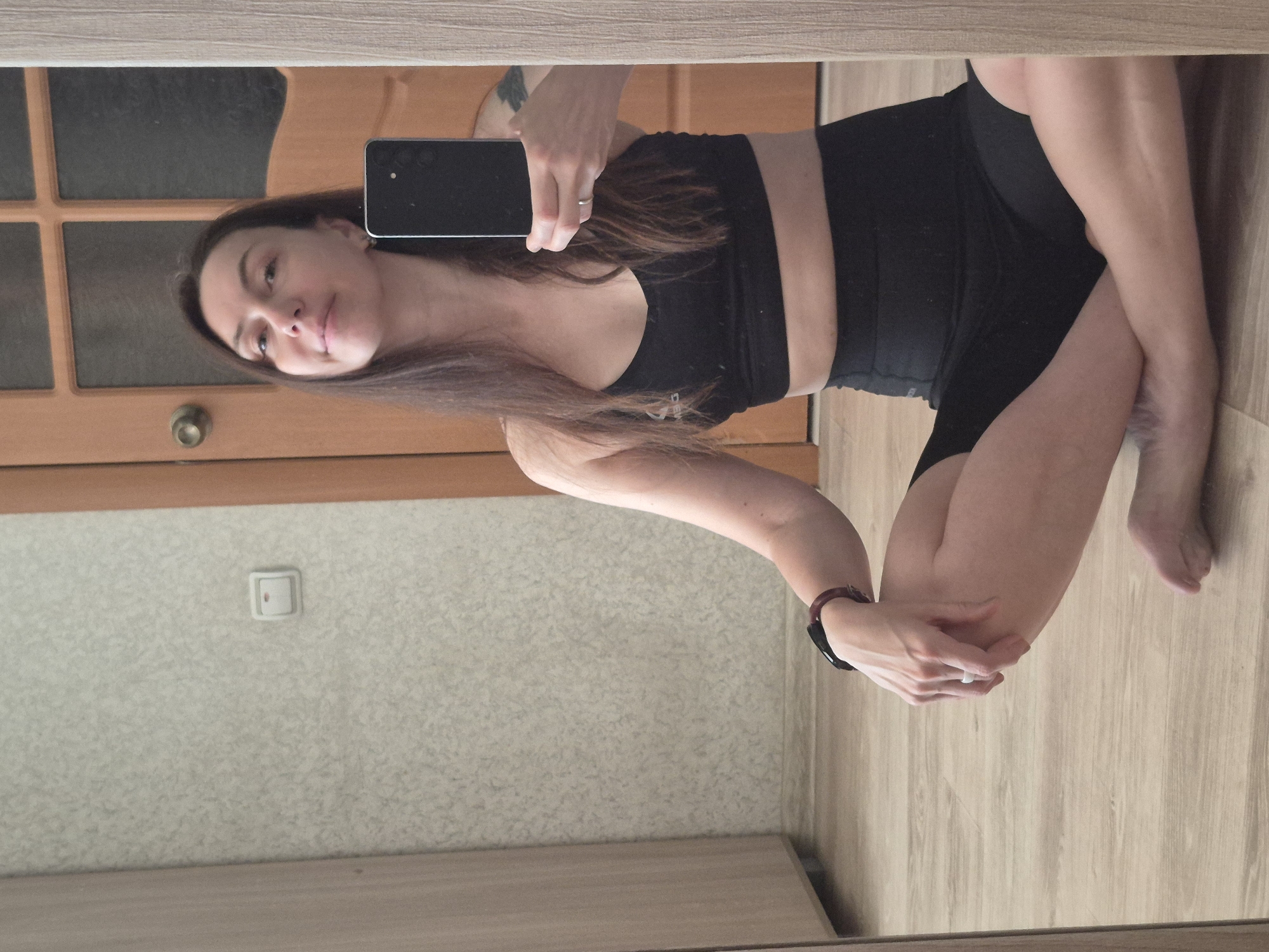 A woman stands in front of a mirror in an indoor setting. She is wearing a black sports bra and black shorts. She holds a smartphone in her left hand, presumably to take a selfie. She has brown hair and is wearing a watch on her left wrist and a ring on the ring finger of her left hand. The background includes a wooden door with glass panels, a light switch on the wall, and wood-patterned flooring. The location appears to be inside a room, potentially an apartment or house.