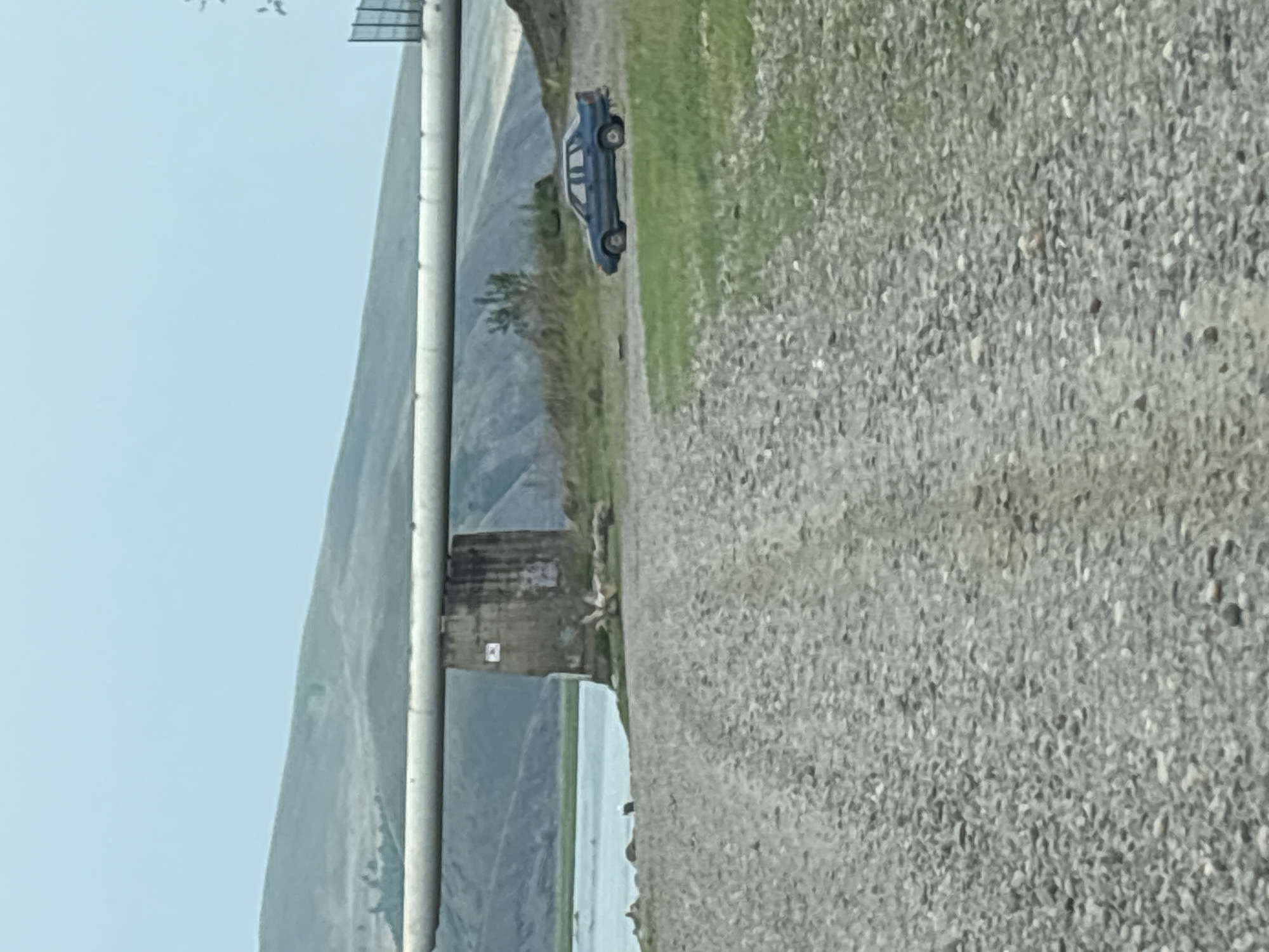 The image depicts a rural landscape scene near Chernogorsk, Russia. In the foreground, a gravel road stretches towards the viewer. In the middle ground, a large pipe, supported by a concrete pillar, traverses the scene horizontally. To the right of the pillar and pipe, a blue sedan is parked on a grassy area. Beyond the pipe and to the left, a body of water, likely a river or lake, is visible, with hilly terrain in the background under a light blue sky. The overall environment suggests an outdoor, undeveloped area.