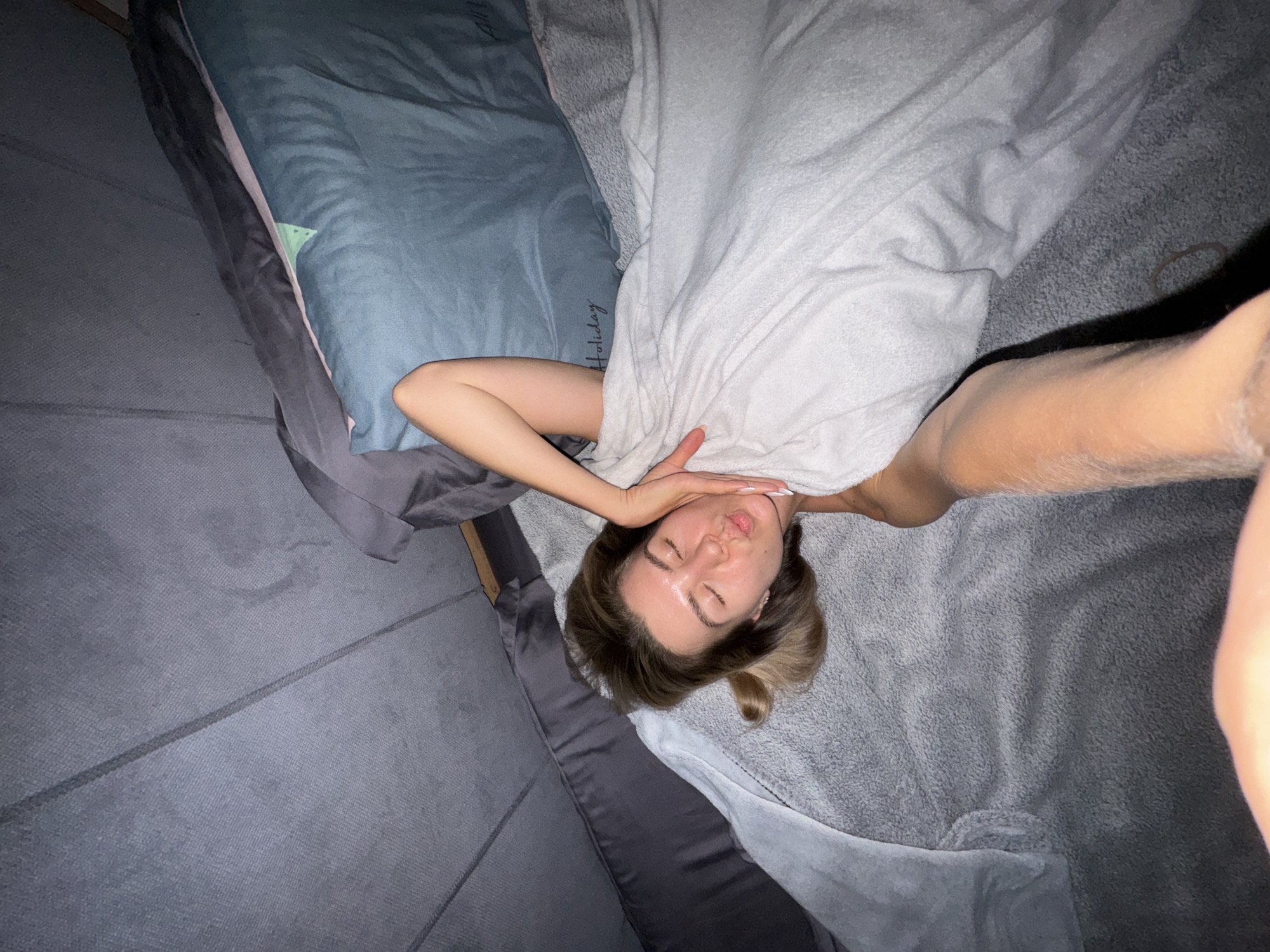 A young woman is lying on a bed with her eyes closed and her lips puckered, as if she is about to kiss someone. Her head is tilted back, and her right hand is resting on her cheek. She is covered by a white blanket, and her left arm is visible. A person's arm is reaching into the frame from the bottom right, touching the blanket. The bed appears to be in a dimly lit room, possibly a bedroom, with a gray couch or mattress in the background. The overall scene suggests an intimate and playful moment.