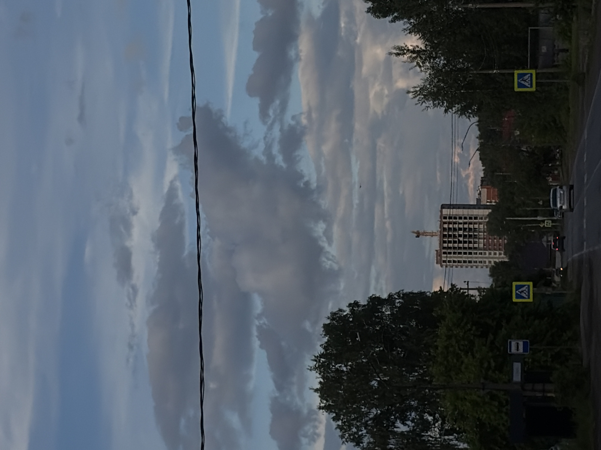 The image shows a street in Unknown, Unknown, with a tall building under construction in the distance. A construction crane is visible atop the building. In the foreground, there are trees and a power line running vertically along the left side of the image. Two pedestrian crossing signs are visible on the right side of the street. A bus stop sign is also present. A car is driving on the street, moving away from the camera. The sky is cloudy, with varying shades of blue and gray.