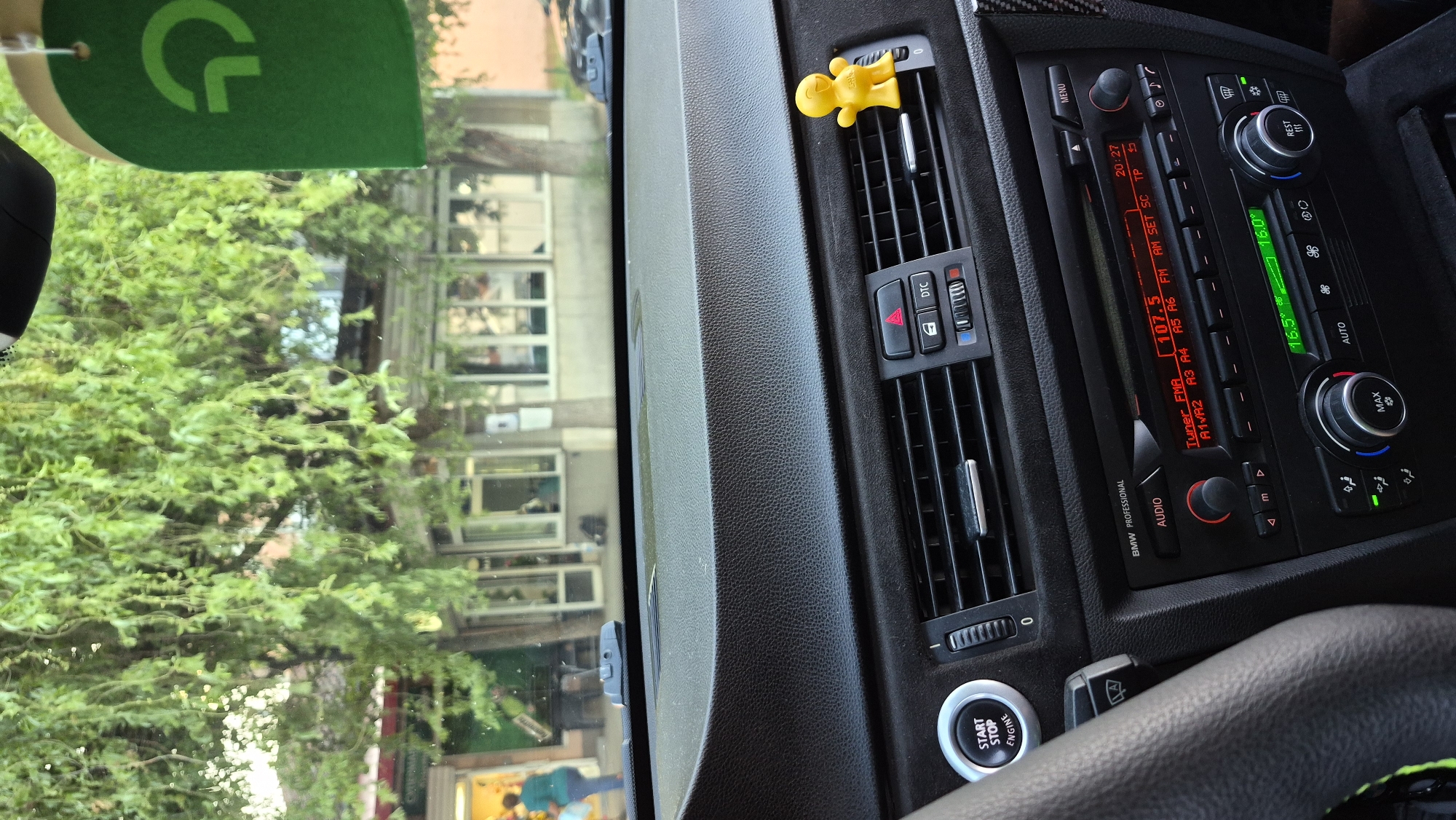 The image shows the interior of a BMW car, looking out through the windshield. The dashboard is visible, with the steering wheel on the left and the radio and climate controls in the center. A yellow bobblehead figure is attached to an air vent on the dashboard. Outside the car, trees with green foliage are visible, along with a building with windows. A person in a teal shirt is walking away from the camera on the sidewalk outside the building. A green air freshener with a logo hangs from the rearview mirror. The time displayed on the car's radio is 20:27.