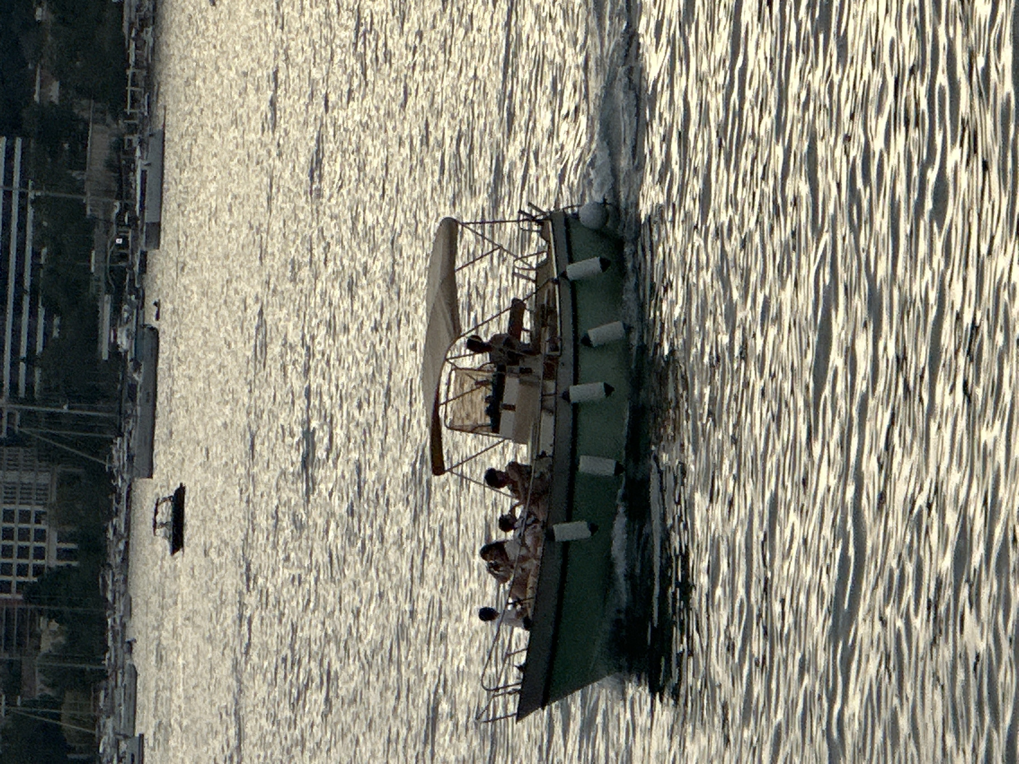A motorboat with a canopy is traveling on the water, leaving a wake. Four individuals are visible on the boat. Two are seated in the front, one standing, and one seated in the rear. The boat is green with white fenders. In the background, there are buildings and other boats moored. The sun is reflecting off the water, creating a shimmering effect. The scene appears to be a harbor or bay in Budva, Montenegro.