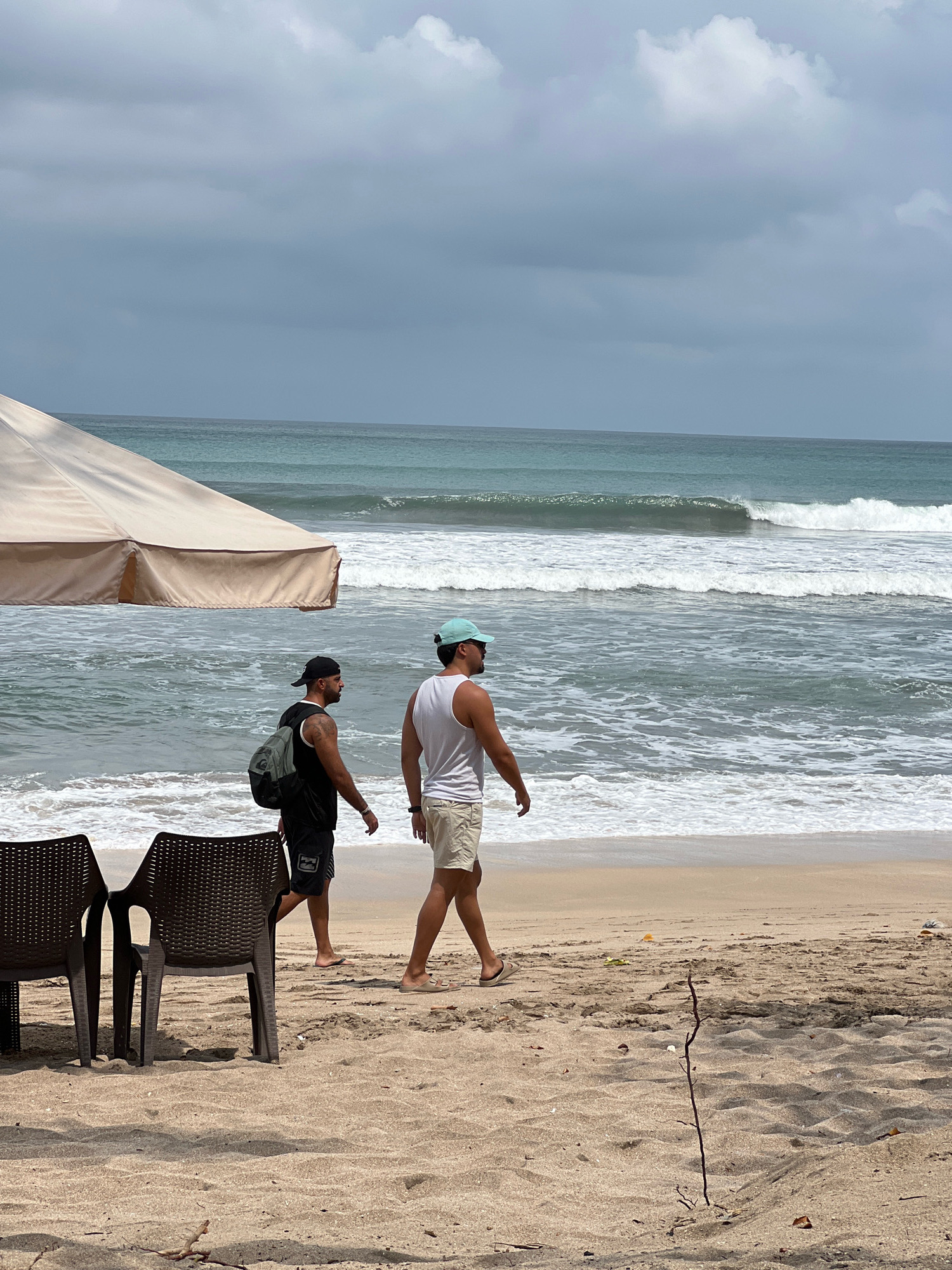 Two men are walking on a sandy beach in Kuta, Indonesia. The man on the left is wearing a black tank top, black shorts, and a black baseball cap, and is carrying a backpack. The man on the right is wearing a white tank top, beige shorts, and a light blue baseball cap. They are walking away from the camera towards the ocean. The ocean has moderate waves and the sky is overcast with some lighter clouds. In the foreground, to the left, are two empty brown plastic chairs and a large beige umbrella. The sand is light brown and scattered with small debris.