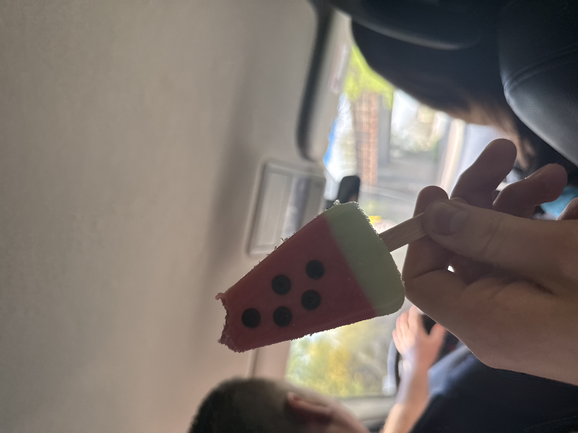 A person inside a vehicle holds a partially eaten watermelon-shaped popsicle. The popsicle is red with black "seeds" and a green "rind." The person's hand is visible holding the popsicle stick. Through the vehicle window, a blurred outdoor scene is visible, suggestive of a neighborhood with buildings and greenery. The location in Golemo Selo, Bulgaria, cannot be verified as there are no recognizable buildings or landmarks in the picture.