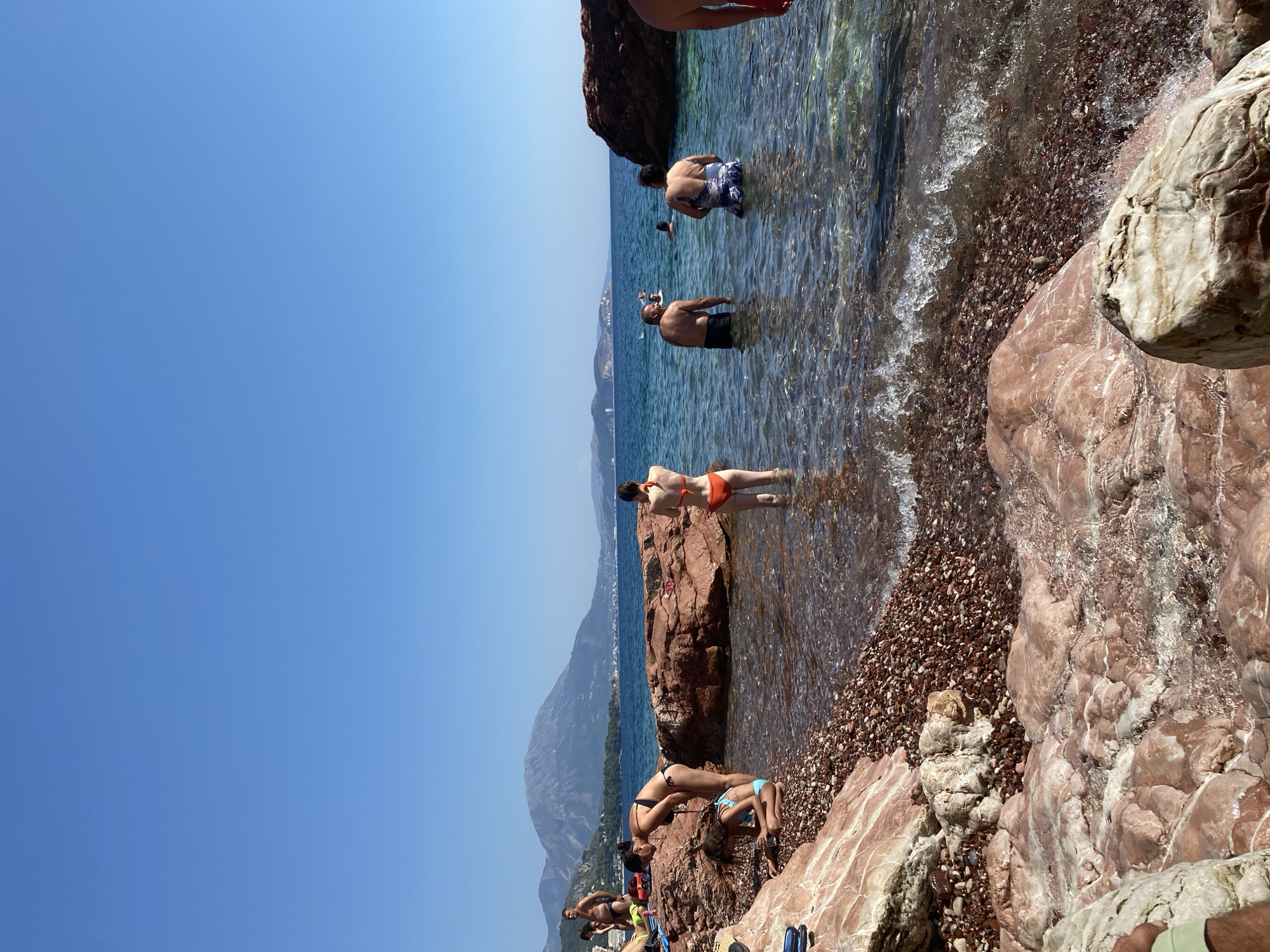 A bright, sunny day is depicted at a rocky beach in Sutomore, Montenegro. The clear, blue sky dominates the upper half of the image, with a distant mountain range visible on the horizon. The foreground is characterized by large, reddish-brown rocks and a shoreline covered in small, dark pebbles. Several people are present, some wading in the shallow, clear water, while others are on the rocky shore.
In the water, from left to right: a woman in an orange bikini stands facing away from the camera. To her right, a man in dark swim trunks is partially submerged, looking towards the shore. Next to him, a woman in a patterned swimsuit is also in the water, facing the same direction. Further out in the water, other individuals are swimming or wading.
On the rocky shore, to the left of the water's edge: a woman in a black bikini is standing and looking down at a young girl in a blue bikini who is kneeling and examining something on the pebbles. Further to the left and slightly behind them, a man in dark swim trunks is standing. Several other people are visible further back on the shore, some lying down.
The water is calm with gentle ripples and small waves lapping at the shore. The rocky formations, both in the water and on land, are a prominent feature of the scene. The overall atmosphere is one of a leisurely day at the beach.