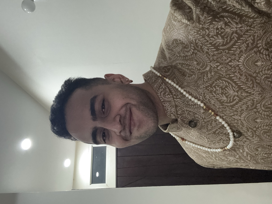 A man with short, dark hair poses in traditional attire. He wears an ornate, gold-colored patterned jacket and a pearl necklace with gold accents. The man looks slightly off-center, smiling at the camera. The background includes a dimly lit room with a closed window and a portion of the ceiling.