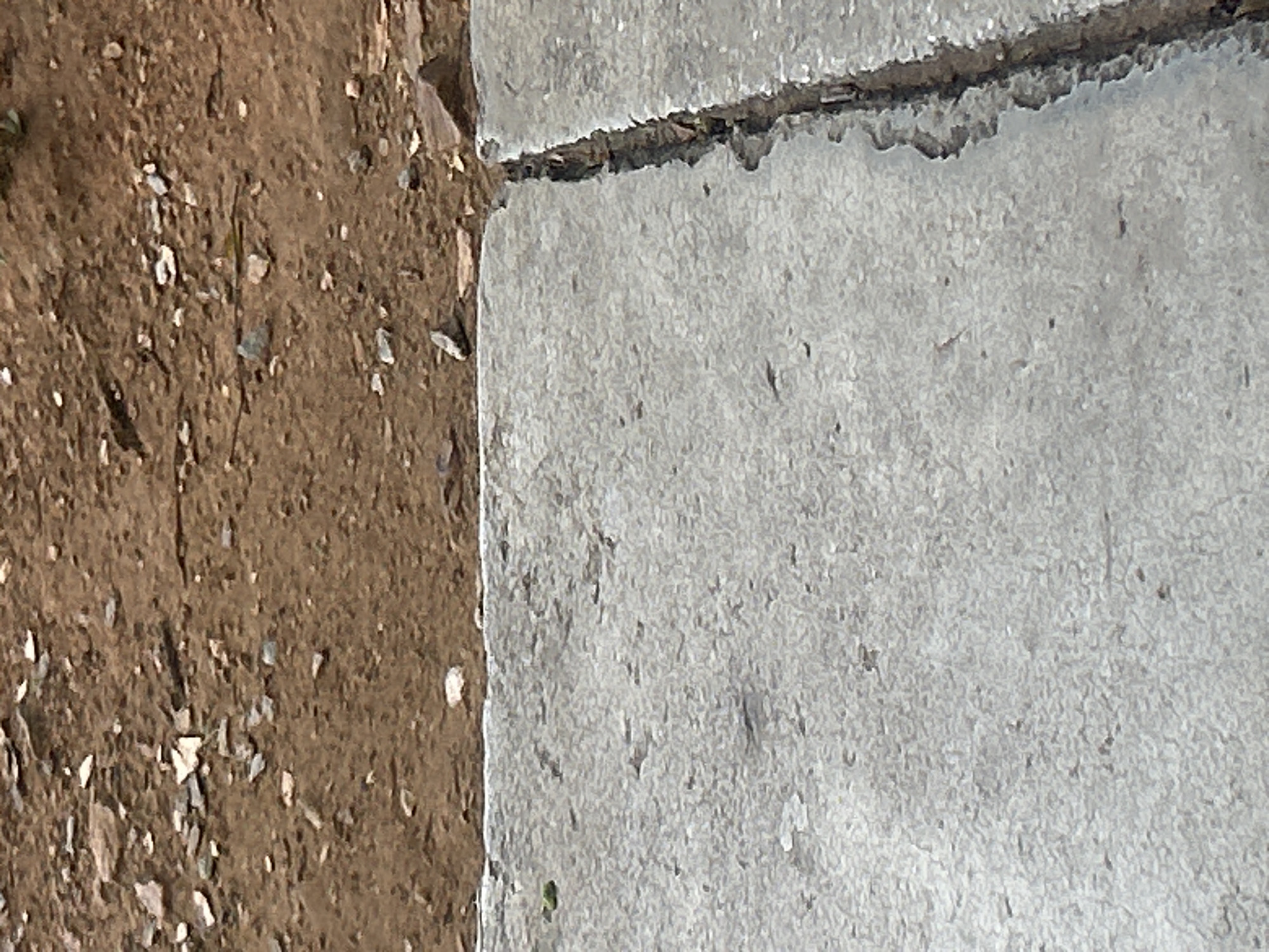 The image shows a cracked concrete surface adjacent to a dirt and gravel area. The concrete appears weathered and is divided by a deep, jagged crack. The dirt area is composed of brown soil mixed with small stones and debris. The scene is captured from a close-up, ground-level perspective. There are no people or specific objects of interest visible beyond the textures of the ground. The location is not discernable from the visual information provided.