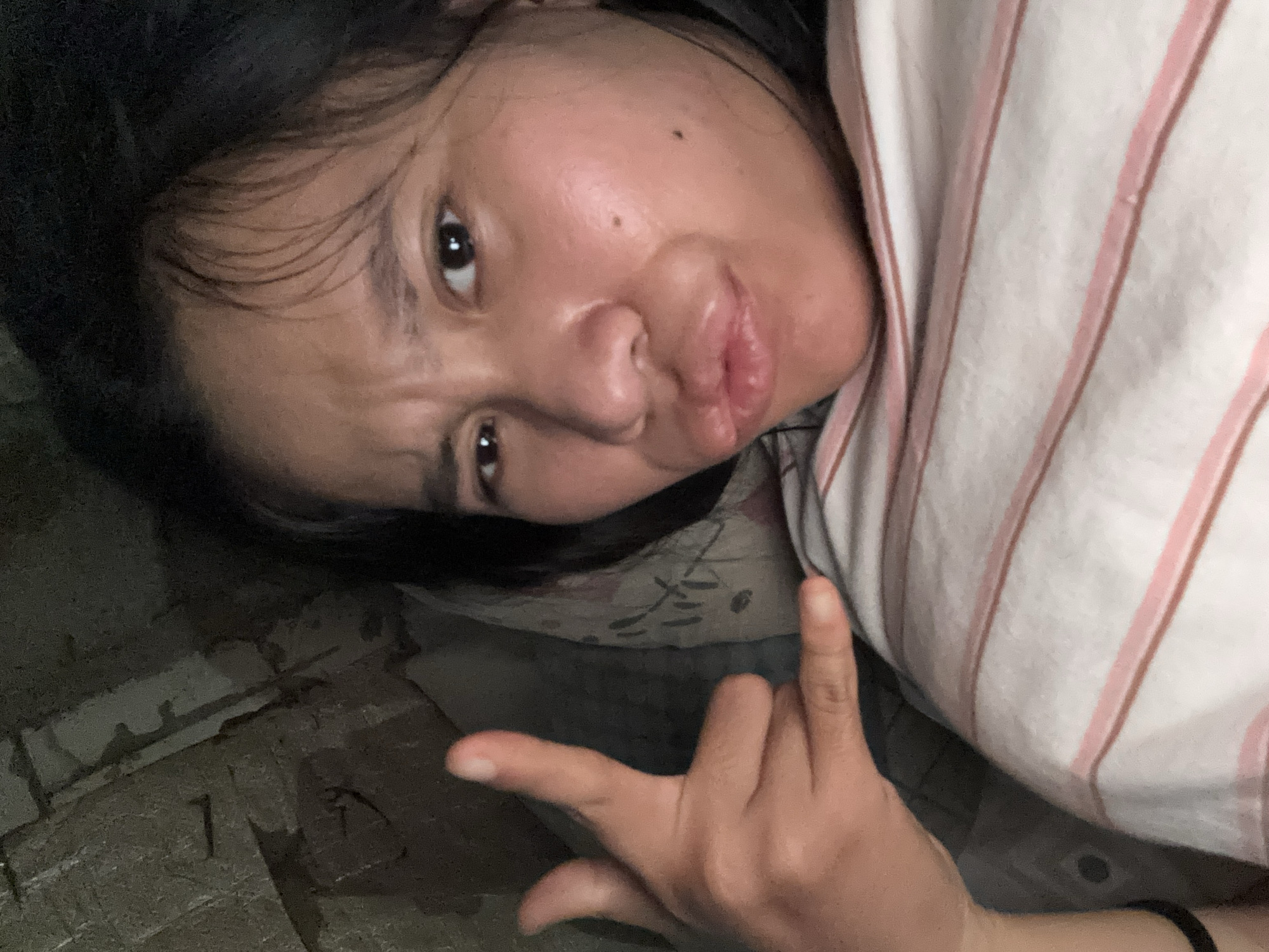 In the city of East Jakarta, Indonesia, a young woman is pictured lying down, seemingly in a bed or on a soft surface, as indicated by the presence of a pillow. She is looking directly at the camera with a playful expression, puckering her lips and raising her eyebrows in a somewhat quirky manner. Her left hand is raised, forming a gesture that could be interpreted as a "shaka" sign or a playful imitation of holding a phone to her ear, with her thumb and pinky extended. She is wearing a striped shirt with thin pink and white stripes. The background is dark and appears to be a wall with textured, possibly concrete, surfaces, hinting at an indoor environment that might not be brightly lit or elaborately decorated. The lighting suggests an indoor setting, possibly at night or in a dimly lit room.