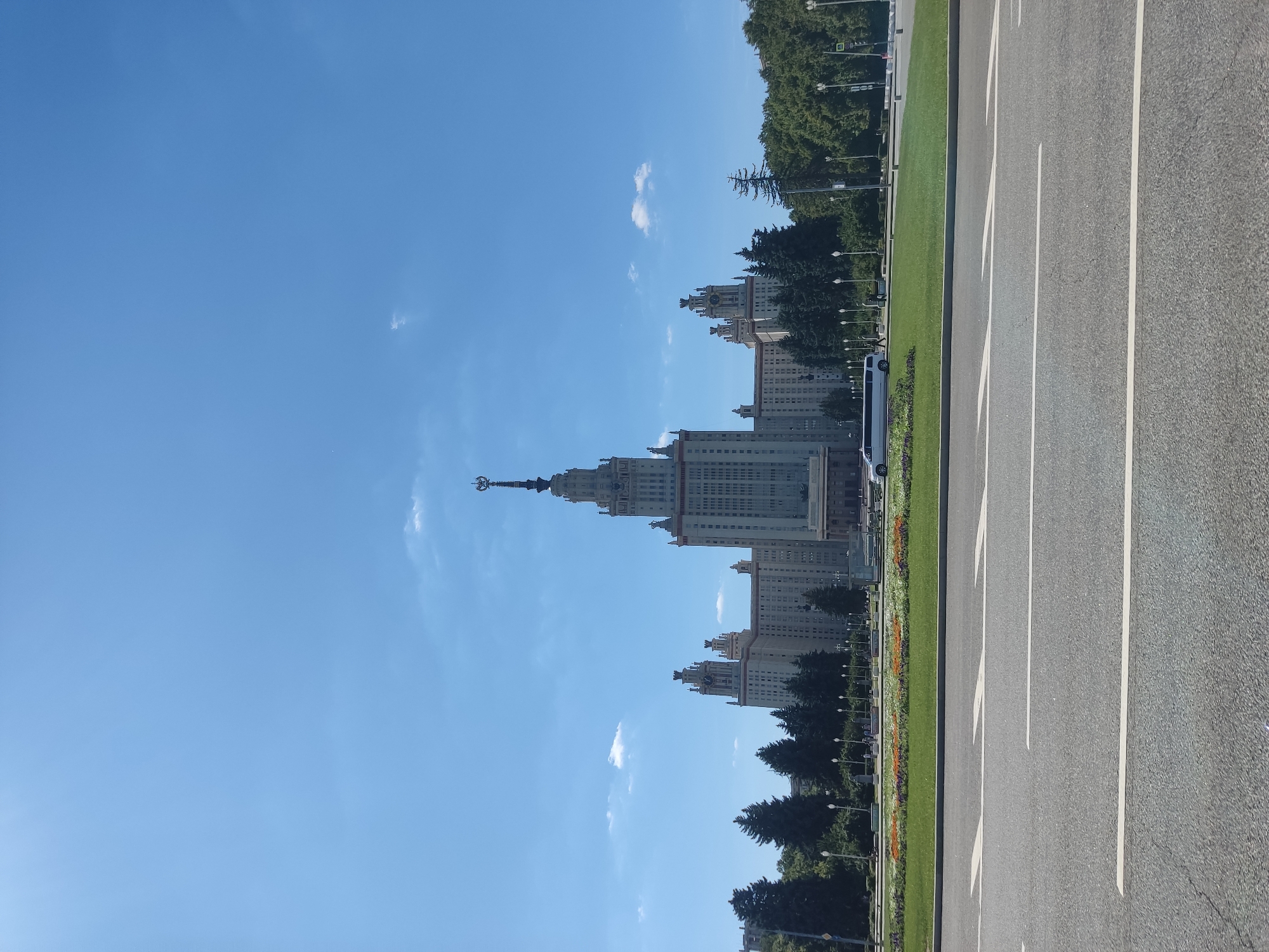 The image depicts a frontal view of a grand, Stalinist-era skyscraper, likely a university building, under a clear blue sky with scattered white clouds. The building is the main subject, towering over lush green trees and a manicured lawn with a flowerbed. A white limousine is parked on the street in front of the building, with a few pedestrians visible in the distance. The foreground shows a portion of an asphalt road with white lane markings. The building's architecture is characterized by its imposing facade, multiple tiers, and a prominent spire at the top. This is likely the Moscow State University, a prominent landmark in the city of Moscow, Russia.