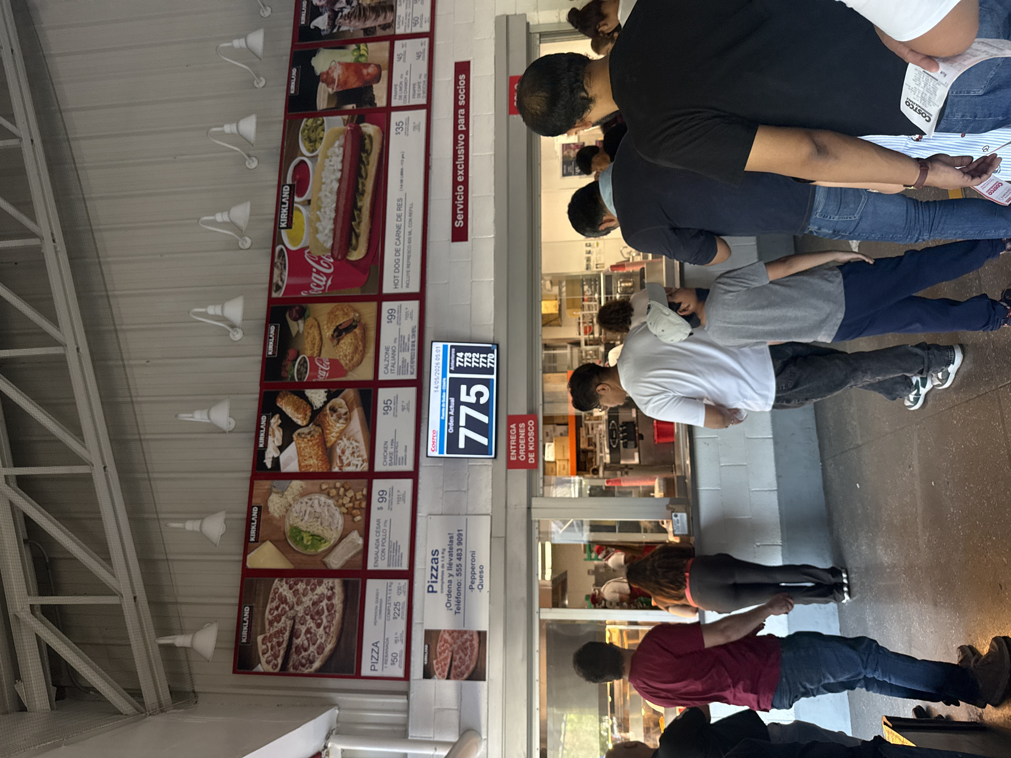 The image depicts a scene at the food court of a Costco store in Mexico City, Mexico. A line of people is waiting to pick up their orders at the kiosk. A digital display shows the order number 775, indicating that it's ready for pickup. The Costco menu is displayed on the wall with photos and prices of items like pizza, hot dogs, salads, and calzones. A sign indicates that the service is exclusive to members. Several white, cone-shaped sconces are affixed to the wall along the menu.