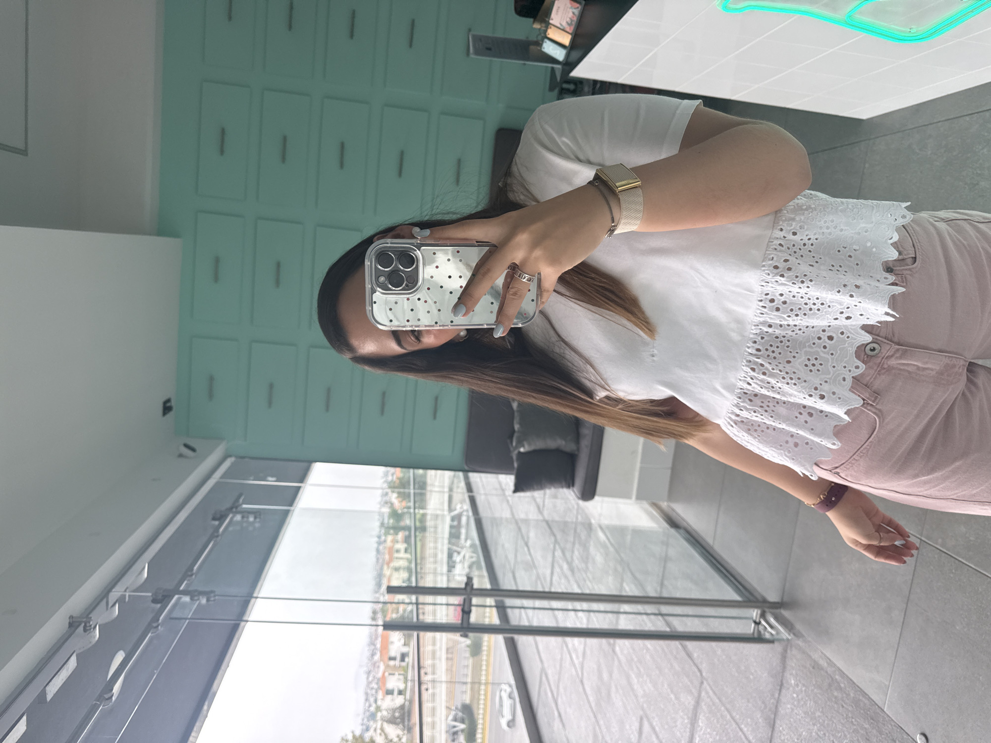 A young woman is taking a mirror selfie in what appears to be a modern apartment or commercial space. She is holding a smartphone with a transparent case, with her face partially obscured by the device. She wears a white top with a lace trim, pink pants, and jewelry on her wrist and fingers.
The location seems to be a high-rise building, as a cityscape is visible through large windows in the background. The buildings and infrastructure suggest an urban setting. The indoor environment features modern design elements like a built-in cabinet with multiple drawers and a light-colored countertop. A grey seating area with throw pillows is visible beyond the glass.
Specific geolocation in San Bernardino Tlaxcalancingo, Mexico cannot be determined with the given information. Further contextual information or landmarks are required for a specific geographical assignment.