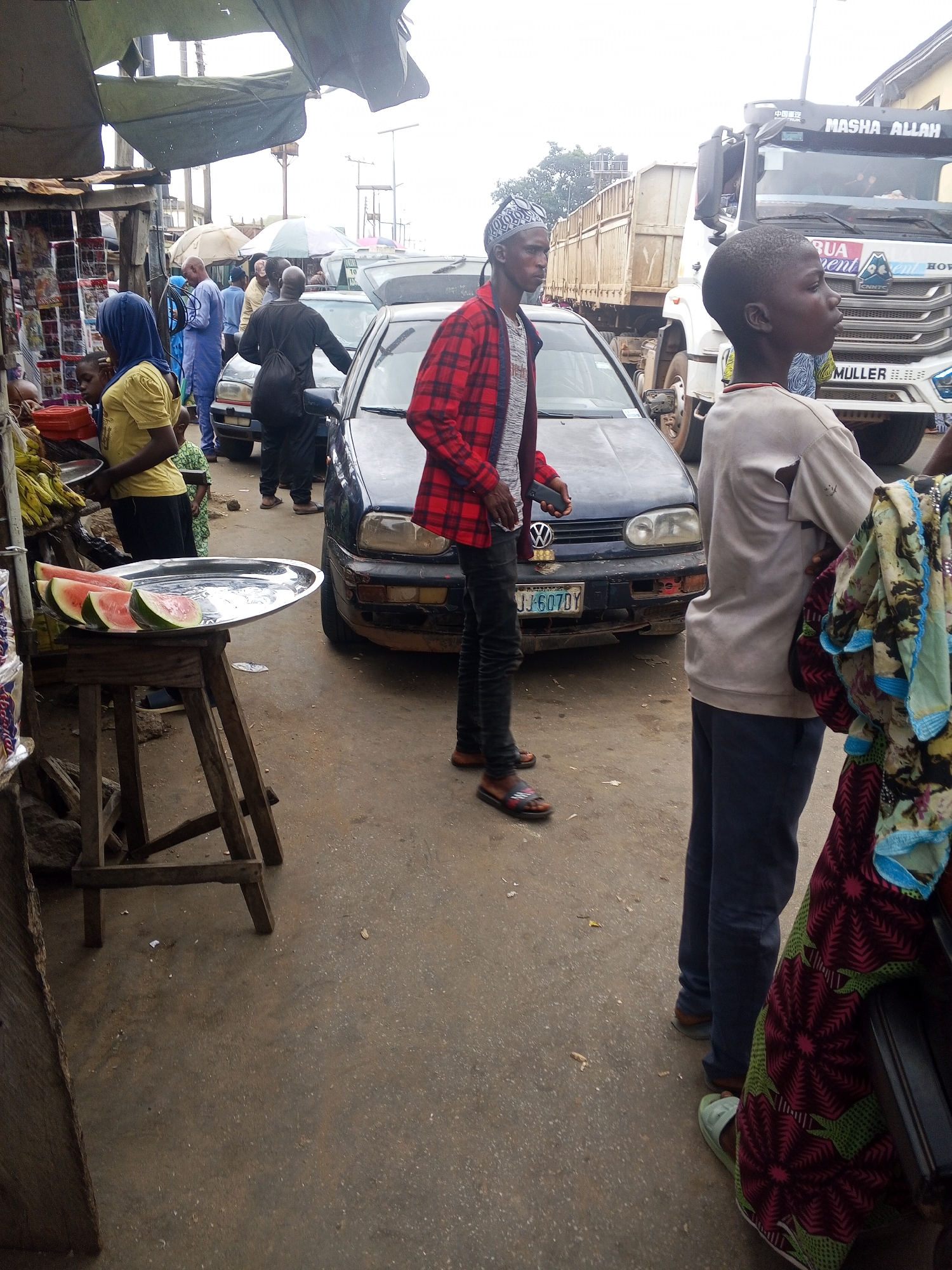 This is a street scene in Okene, Nigeria, likely a marketplace, with several individuals present amidst parked vehicles and market stalls. In the foreground, a person wearing a red and black plaid shirt and dark pants stands beside a dark blue Volkswagen Golf. To the right, a young person wearing a light-colored t-shirt and dark pants is visible, partially obscured by a woman in a brightly patterned fabric. A metal tray with several slices of watermelon is displayed on a wooden stand.
Further in the background, several other people are seen. A man in a dark outfit with a bag slung over his shoulder is near the Volkswagen. Another man in a blue robe stands near a car. A large truck with "MASHA ALLAH" visible on its windshield is parked on the right side of the street. Market stalls with awnings are visible on the left, displaying various goods. The ground is paved with concrete, and the overall atmosphere suggests a busy, active urban environment.