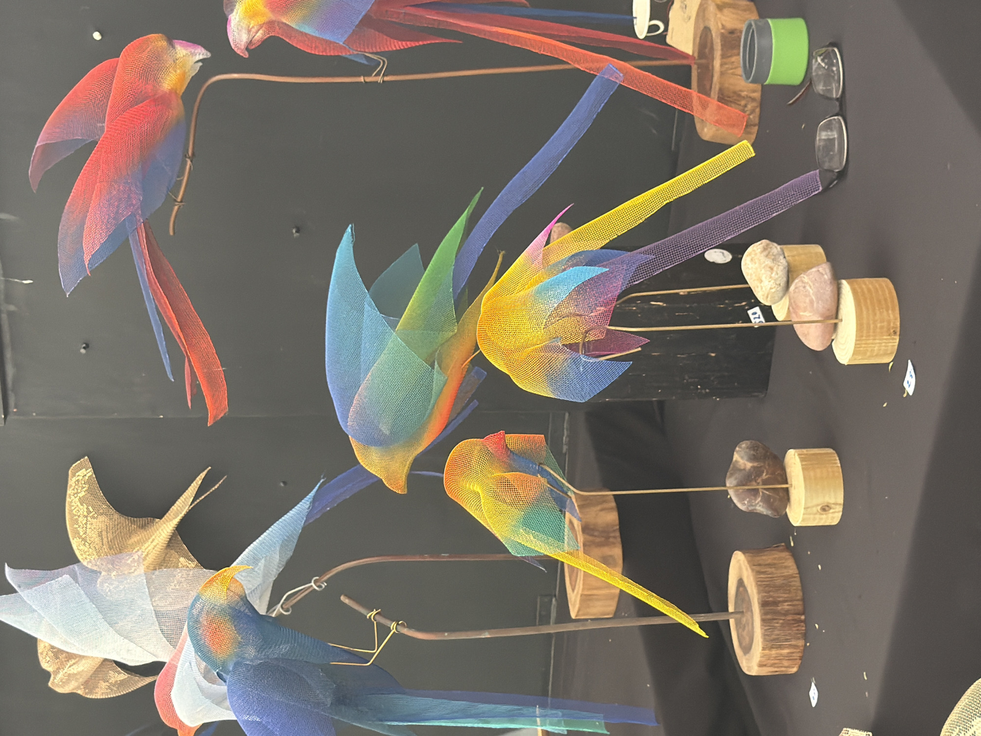 The image displays an artistic arrangement of multiple bird sculptures crafted from vibrant, translucent mesh fabric. The birds are diverse in shape and size, featuring gradient color schemes including reds, blues, yellows, greens, and purples, giving them a rainbow-like appearance. Some larger birds resemble macaws with long tails, while others are smaller and more abstract in form.
Each sculpture is mounted on a thin metal rod or wire, which in turn is secured into a base. These bases vary, consisting of natural wooden log cross-sections, and combinations of wooden logs topped with smooth, polished stones of different colors like grey, pink, and brown.
The sculptures are displayed on a dark, flat surface, creating a stark contrast that highlights their bright colors and delicate construction. The background is uniformly dark, suggesting an indoor setting such as an art exhibition, craft fair, or retail display.
Visible alongside the sculptures are a few additional items: a small green and grey cylindrical object, two pairs of eyeglasses, and a white ceramic mug resting on a wooden base. One of the wooden bases has a small white tag with black text, partially visible, which appears to indicate "20" or "$20", suggesting a price tag. Another similar tag is partially obscured. No people are visible in the image. The overall scene conveys a sense of creative craftsmanship and artistic presentation.