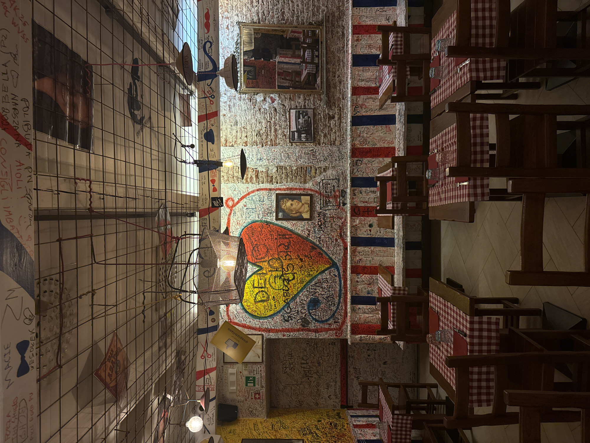 The image depicts the interior of a restaurant, possibly located in Milan, Italy, based on the presence of Italian flags and writing on the walls. The restaurant's walls are covered in graffiti, signatures, and drawings, including a large heart-shaped design in the center. The lighting is dim, and the tables are set with red and white checkered tablecloths. There are chairs arranged around the tables, and the overall atmosphere appears informal and artistic. A wire mesh structure runs along one wall, adding to the eclectic decor. Several framed pictures and mirrors are also visible on the walls.