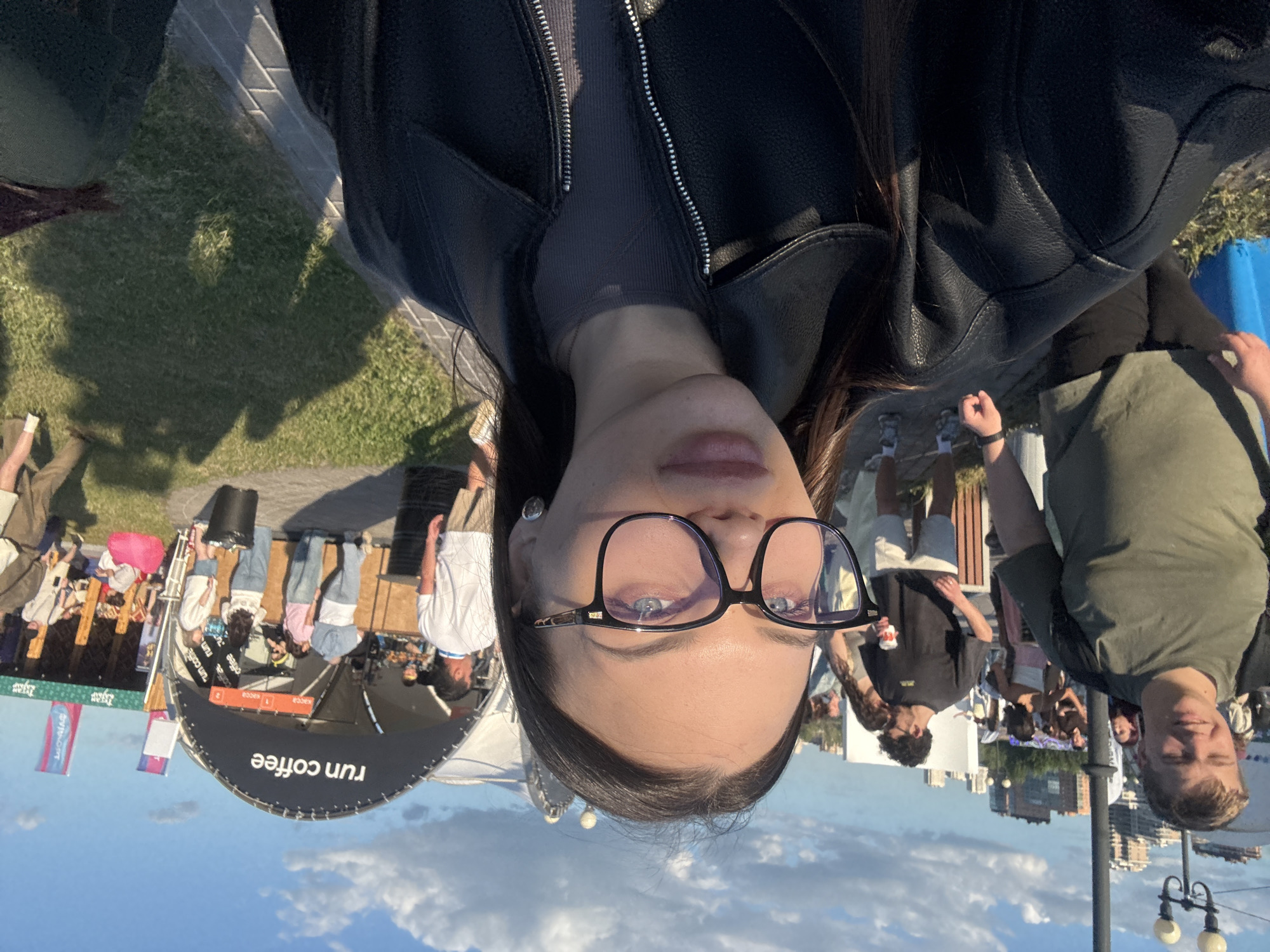 The image presents an outdoor event scene in Kazan, Russia, under late afternoon or early evening sunlight, indicated by warm light and long shadows. A young woman occupies the foreground, taking a selfie. She has dark hair and wears black-framed glasses with a prominent blue reflection on the lenses, largely obscuring her eyes, though a light blue iris is visible in her left eye. She wears a black leather jacket over a dark grey ribbed top with a zipper. A heart-shaped earring is visible on her left ear.
In the mid-ground to the left, a young man with light hair, wearing a green t-shirt and a dark backpack, walks towards the viewer with a slight smile. Behind him, a streetlight pole and several other individuals are visible. The far background features a city skyline with multiple buildings against a blue sky with scattered white clouds.
To the right of the frame's center, a prominent, dark-colored arched tent structure with "run coffee" printed in white serves as a coffee stand. Several people are gathered around the stand, appearing to order or wait. A black trash can is situated near the stand. Further to the right, another tent structure is partially visible, along with various individuals on a grassy area, some sitting on pink bean bags. The ground consists of paved pathways and well-maintained grass.