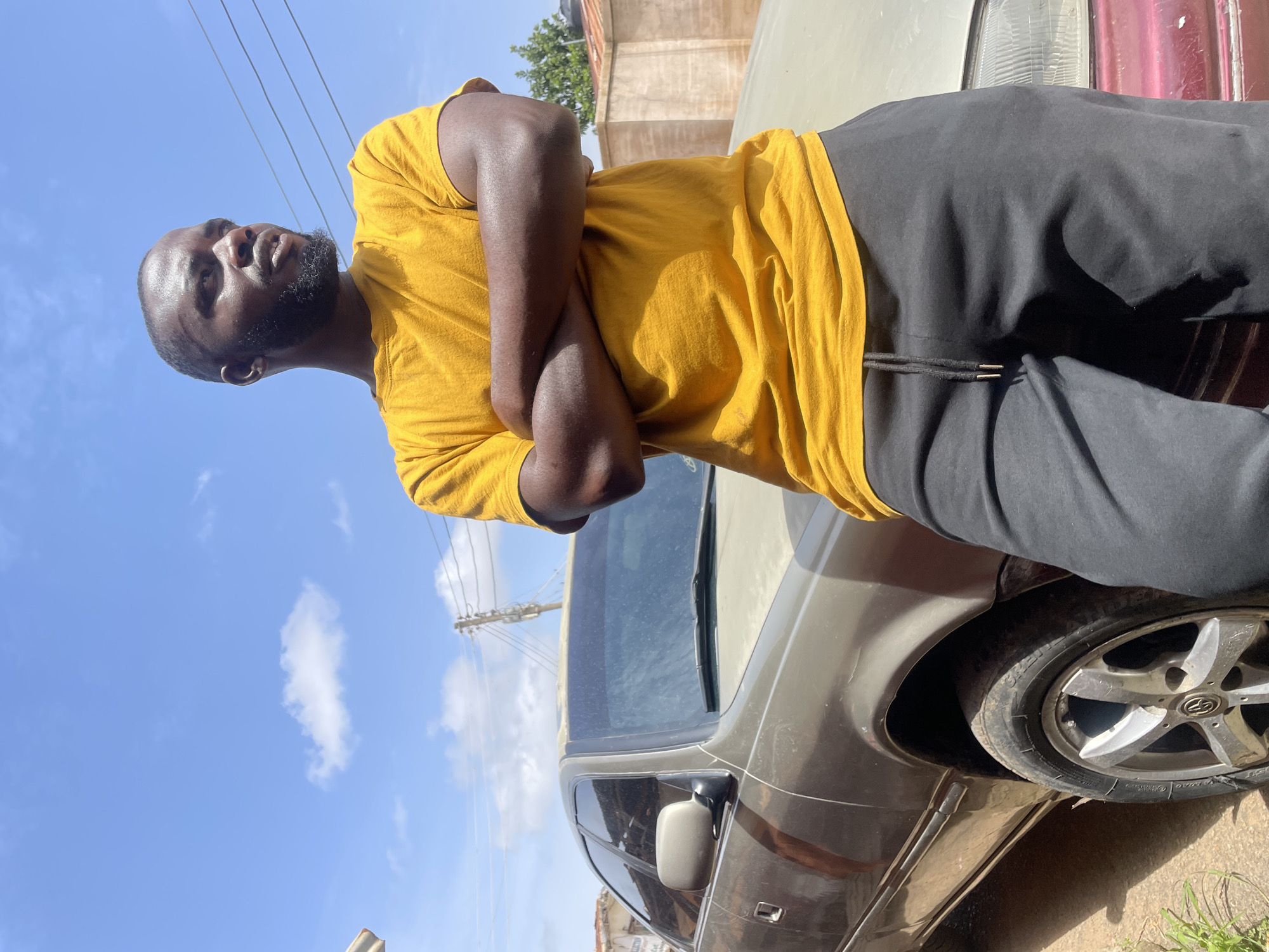 The image depicts a dark-skinned man standing next to a car. He is wearing a yellow t-shirt and black pants. His arms are crossed. The car is gray or silver. The background includes a blue sky with some clouds and power lines. The location is likely in Jos, Nigeria.