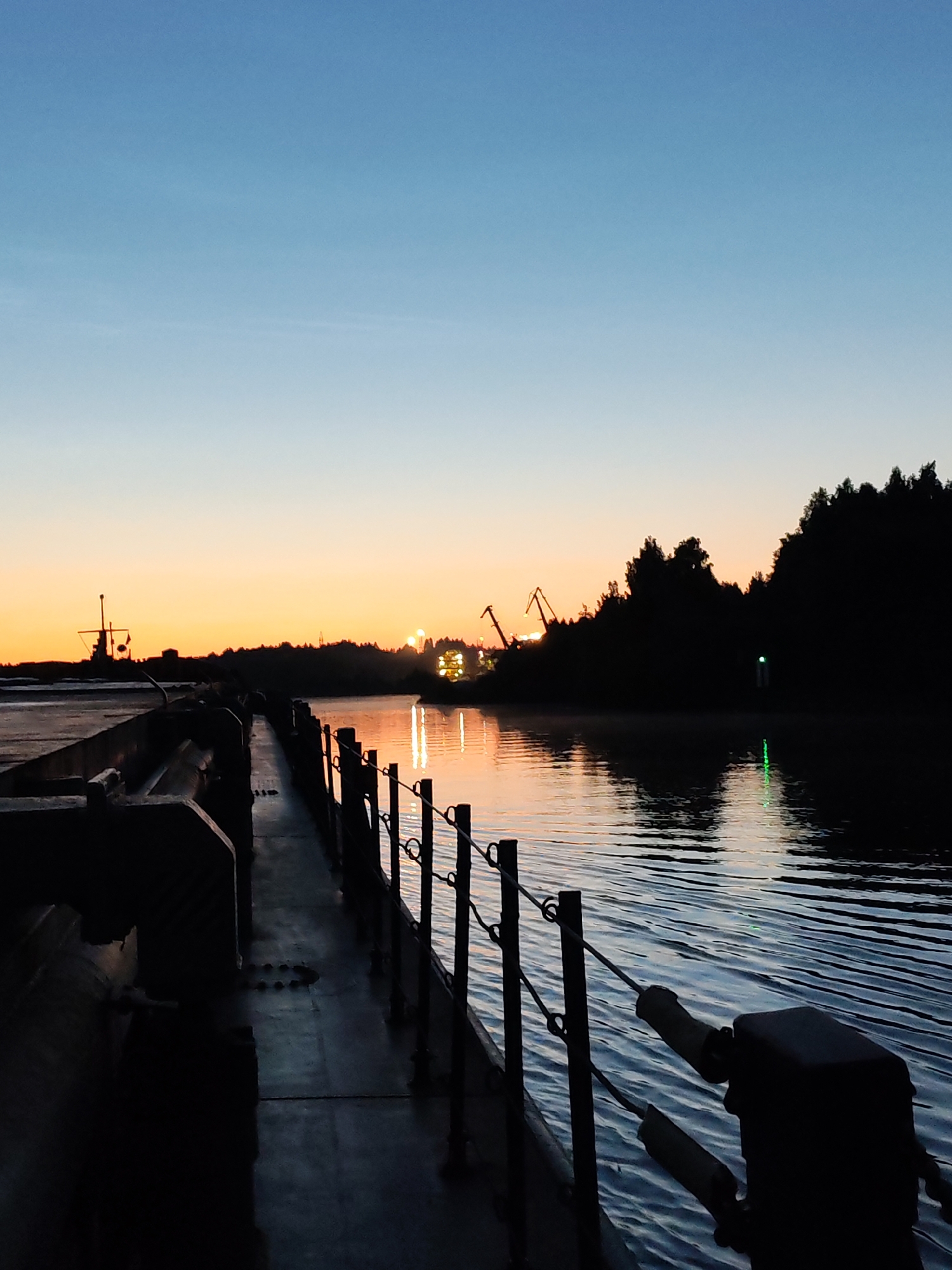 A low-angle, silhouette view of a dock at dawn with a clear, gradient sky transitioning from deep blue to orange. The foreground features the dark, industrial structure of a dock or pier, with a railing and cleat visible on the right. A body of water, reflecting the sky's colors and distant lights, stretches to the right. Silhouetted against the horizon are trees, industrial cranes, and a ship. In the distance, the faint glow of lights suggests a town or industrial area. The overall scene evokes a sense of quiet industrial activity at the beginning of a new day.