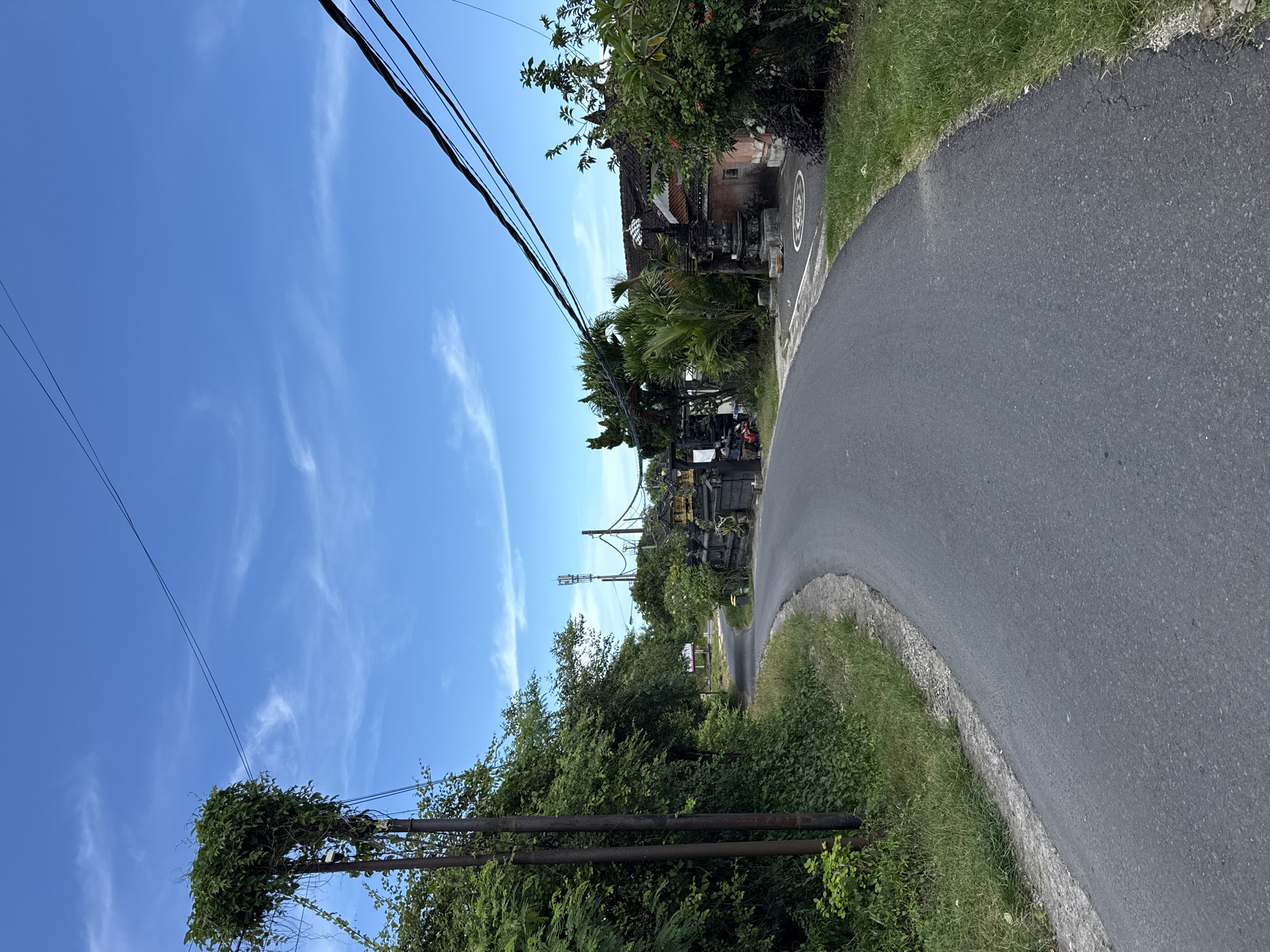 The image depicts a narrow asphalt road curving through a landscape in the city of Kutuh, Indonesia. The sky is a clear blue with faint wispy clouds. Power lines stretch across the upper portion of the frame. On the right side of the road, lush greenery and a traditional Balinese building are visible. The road surface appears well-maintained. On the left, overgrown foliage covers utility poles, adding to the naturalistic aesthetic. The perspective suggests the photo was taken at ground level. There are no visible people or other subjects clearly identifiable in the scene.