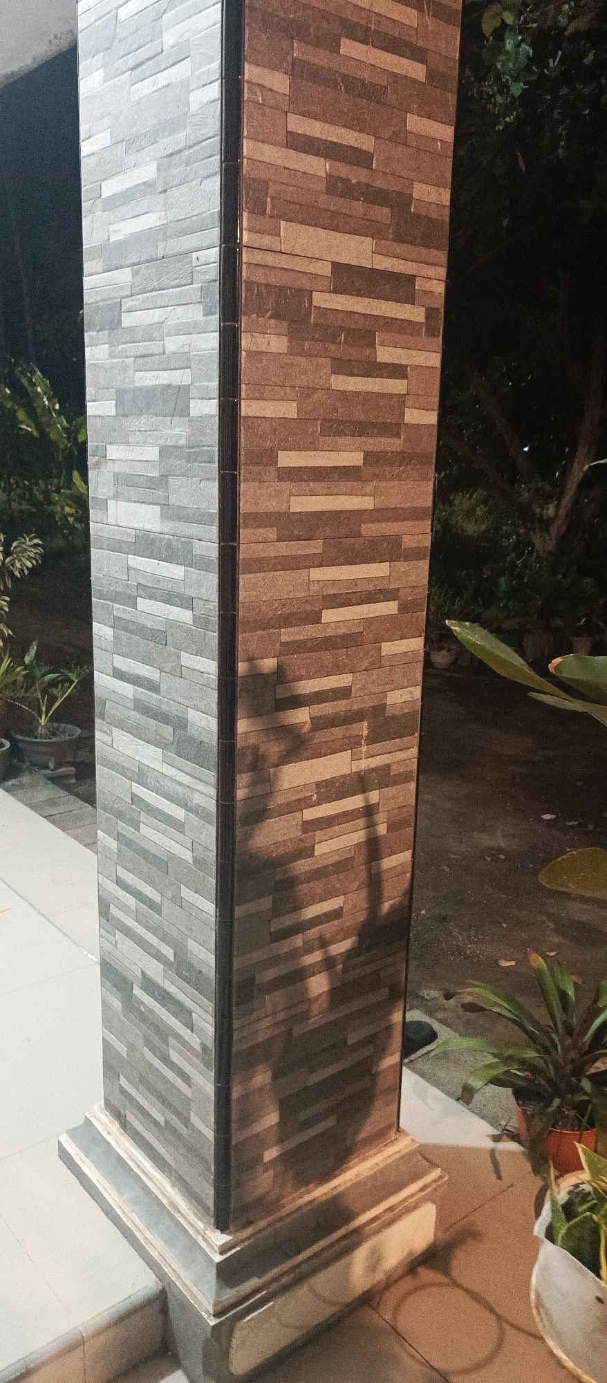 The image shows a close-up view of a pillar on the exterior of a building, likely a residential property, in Arjasa, Indonesia. The pillar is rectangular and covered in stacked, rectangular tiles. The left side of the pillar features gray and white tiles, while the right side is adorned with brown and beige tiles. A dark vertical seam separates the two sections of tiles. The pillar stands on a multi-layered, stepped base. Shadows of foliage and possibly a person are cast onto the brown and beige side of the pillar, suggesting it is daytime and there are plants and trees nearby. In the background, out-of-focus foliage and dark areas indicate an outdoor setting at night or in shadow. The ground appears to be concrete or paving stones. There are no people or identifiable actions depicted.