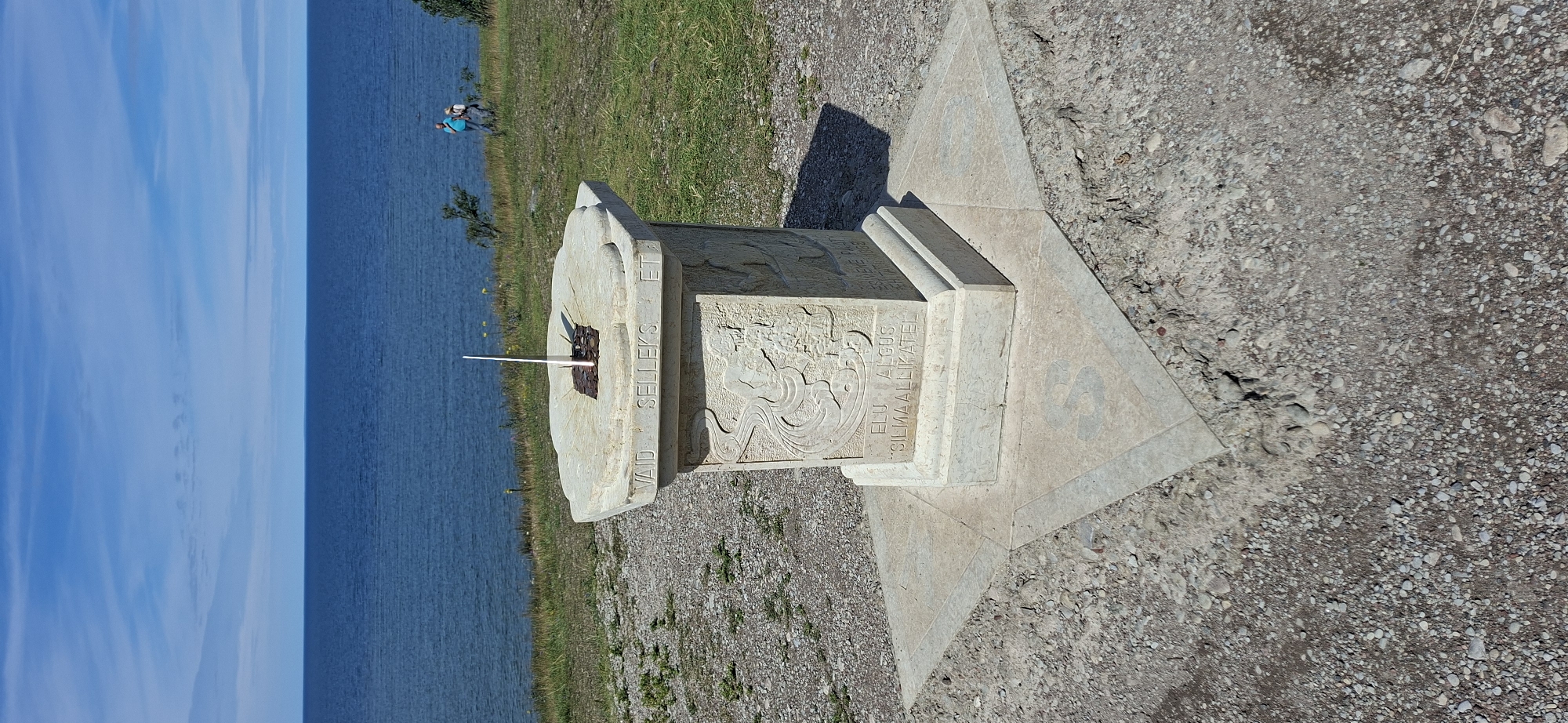 A sundial monument stands on a star-shaped base on a pebbly ground. The sundial face is engraved with Roman numerals and has a gnomon. The stone pedestal of the sundial is decorated with reliefs. In the background, the blue sea meets the horizon under a blue sky with scattered clouds. Two people are visible in the distance, standing on a grassy slope near the sea. The location is identified as Panga, Estonia.