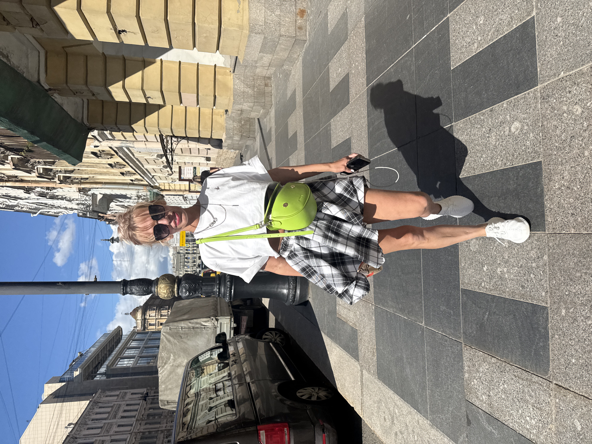 A woman with blonde hair, wearing sunglasses, a white t-shirt, and a black and white plaid skirt, stands on a city sidewalk. She has a bright green crossbody bag and is holding a smartphone in her right hand. Her white sneakers are visible. The background shows a building with light-colored stonework and architectural details, a black lamppost, and a street with a parked black vehicle. The sky is blue with some clouds. The scene appears to be outdoors during the daytime, likely a sunny day given the strong shadows. The image is taken from a slightly elevated perspective, looking down at the woman. The overall impression is of a casual stroll in an urban environment. The setting suggests a city street in Saint Petersburg, Russia, with its characteristic architecture and cobblestone-like paving.