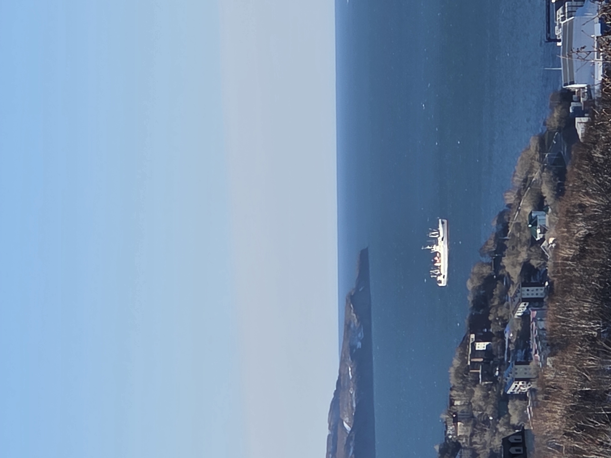 The image shows a body of water adjacent to land. A large ship is visible on the water. Along the land, there are buildings and trees, suggesting a coastal town or city. The sky is clear and light blue. The photo appears to be taken from an elevated viewpoint. The city is unidentified.