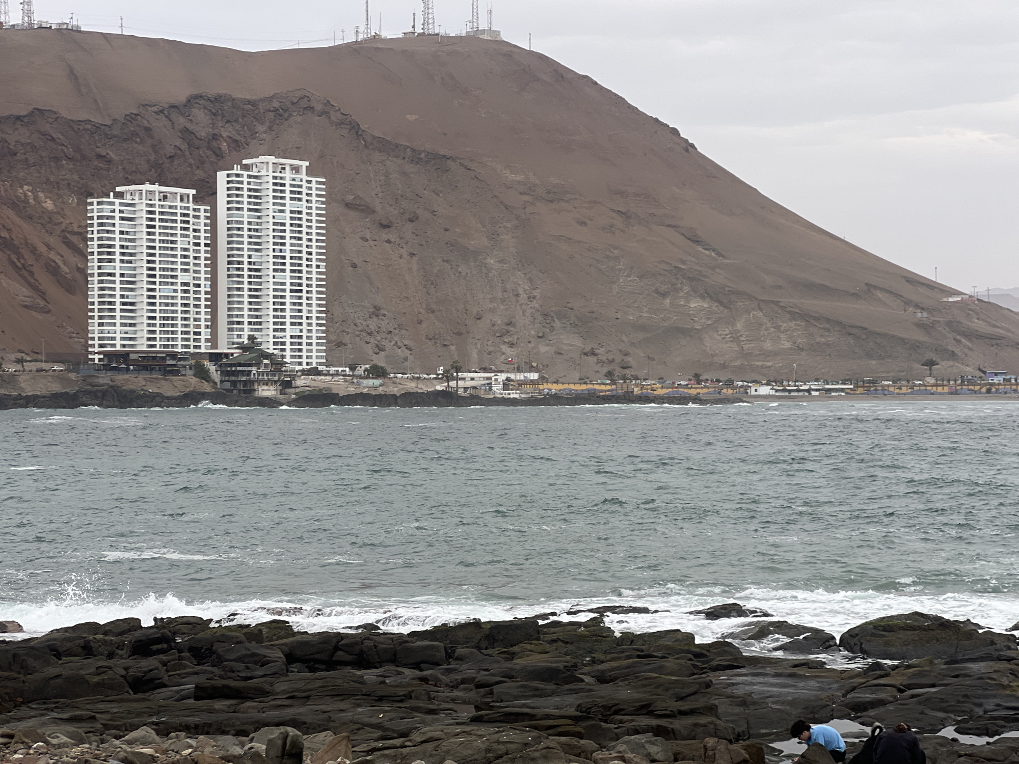 In the city of Arica, Chile, the image captures a coastal scene with a focus on the striking natural and man-made elements. Dominating the background is a large, reddish-brown mountain with rugged textures and sparse vegetation, topped with several communication towers. Nestled against the mountain's base are two identical, tall white apartment buildings with a grid-like pattern of windows. Below these buildings, a line of smaller structures, including houses and businesses, stretches along the coastline. The foreground is characterized by a rocky shoreline with dark, irregular rocks of various sizes. The ocean in the middle ground exhibits choppy, gray-green water with white waves crashing against the rocks. Two individuals are present in the lower right foreground, crouched among the rocks, seemingly engaged in an activity near the water's edge. The sky is overcast and a pale gray.