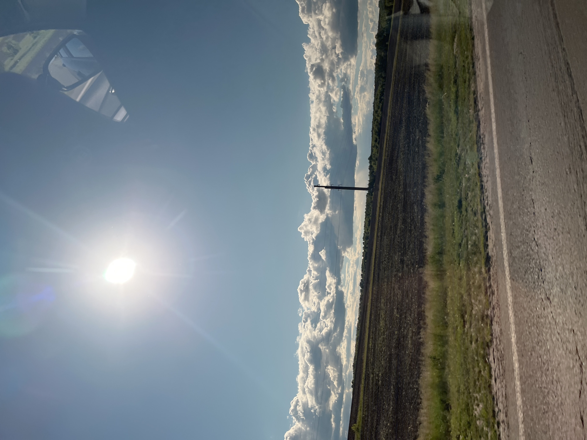 The image shows a view from inside a vehicle, looking out over a rural landscape. The sun is brightly shining in a blue sky, partially obscured by lens flare. A strip of puffy white clouds lines the horizon, bordering a harvested field. A power line is visible between the field and the clouds. A narrow strip of green vegetation runs between the field and an asphalt road. The side view mirror of the vehicle is visible in the upper left corner. The location is unknown.