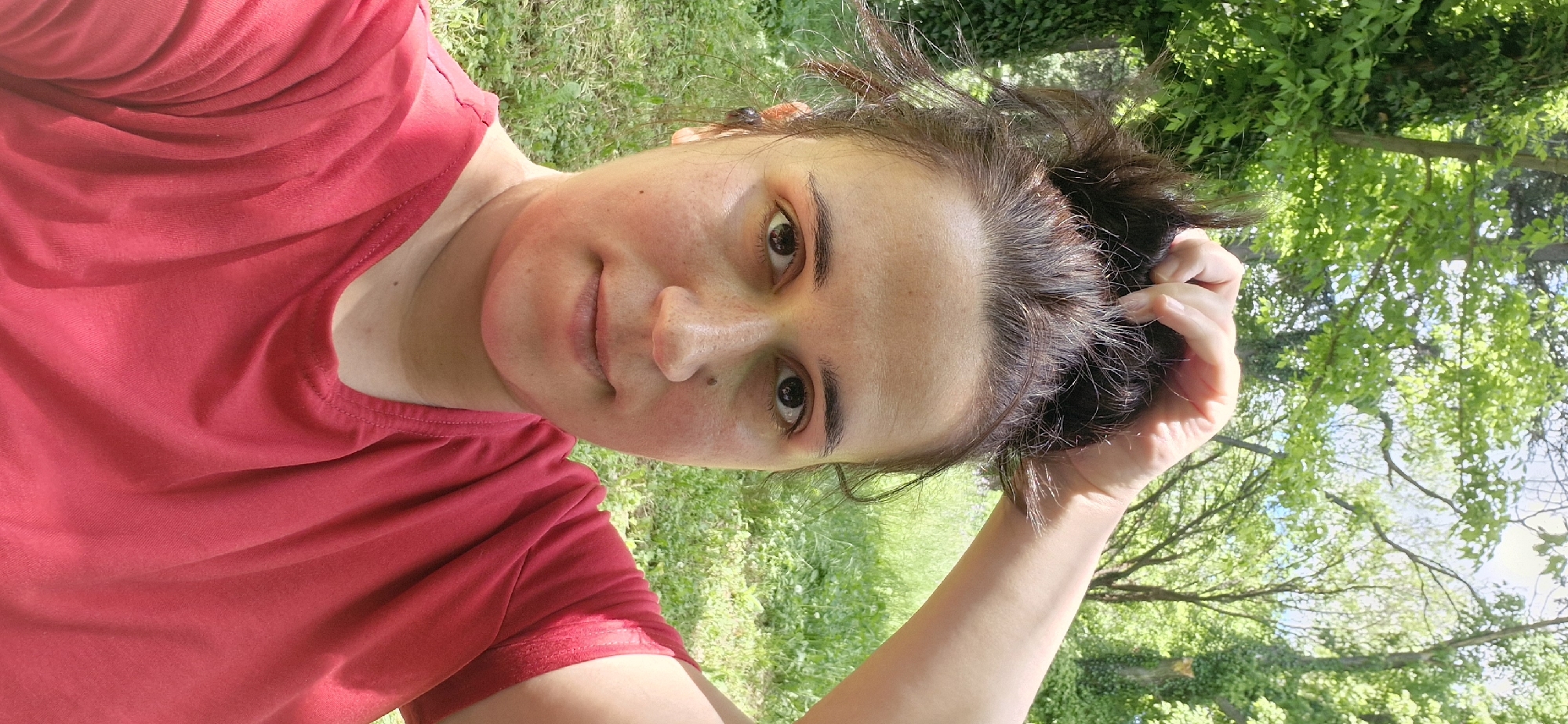 A woman with brown hair and a red shirt stands outside in a green, grassy area with trees. She is holding her hair with her right hand. The location is possibly in or around Stara Zagora, Bulgaria, given the prompt's request.