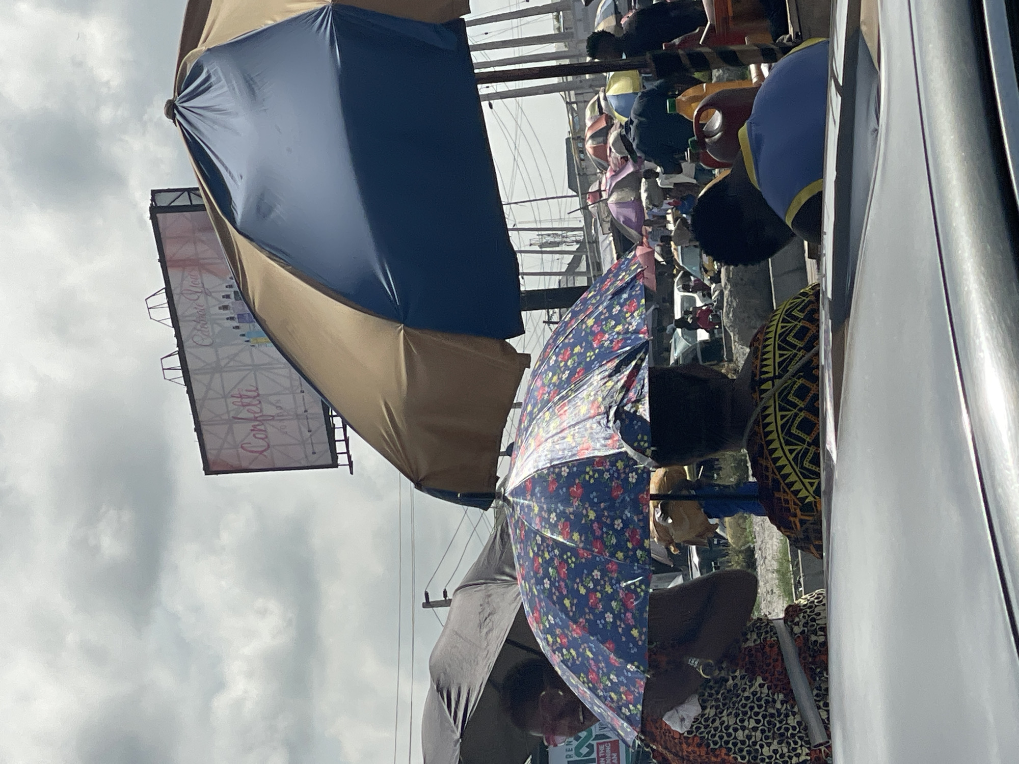 This image captures a vibrant, rotated street scene, likely a market, in Satellite Town, Nigeria, under an overcast sky. The photograph appears to be taken from inside or near a vehicle, looking out onto a busy thoroughfare.
In the prominent foreground, a large, two-toned umbrella, dark blue on top and tan underneath, dominates the left side, angled upwards. Next to it, an open, floral-patterned umbrella with blue, red, and yellow hues provides shade. Below this umbrella, a person is visible, their head and shoulder obscured by the umbrella, wearing clothing with striking, patterned fabric. Another individual in a bright blue top with yellow trim is seen to the right, facing away, close to the side of a white vehicle that frames the right edge of the photo.
The background is filled with the activity of what appears to be a bustling market. Numerous other umbrellas, some striped and multi-colored, are spread throughout, sheltering vendors and shoppers. People are visible amidst the stalls, engaging in daily market routines. Utility poles and a network of overhead wires are noticeable against the cloudy sky, indicating an urban or semi-urban setting.
High above the market, a rectangular billboard stands out against the clouds. The word "Confetti" is clearly visible in a decorative script font on the billboard. Below the floral umbrella, a small green sign with partially visible text like "REN" and "ON THE" can be glimpsed, along with the number "25" on a smaller white sign. The cloudy conditions suggest daytime, possibly a cool or overcast day, conducive to outdoor market activities.