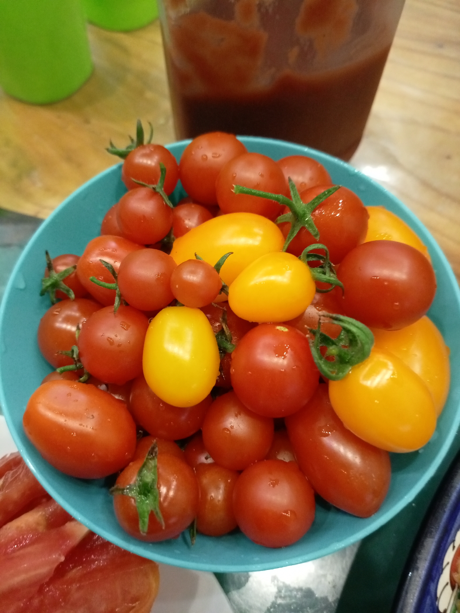 A close-up, overhead shot shows a teal bowl filled with cherry tomatoes. The tomatoes are a mix of red and yellow, with some still attached to their green stems. The bowl is placed on a wooden surface, and in the background, out of focus, are two green cups and a plastic container filled with a dark liquid, likely tomato sauce. Some of the tomatoes have water droplets on them, suggesting they have been recently washed. The lighting is bright and appears to be natural. The scene appears to be an indoor setting, possibly a kitchen or dining area. The location information provided (Березки Снт, Russia) indicates a specific geographical context for this image.