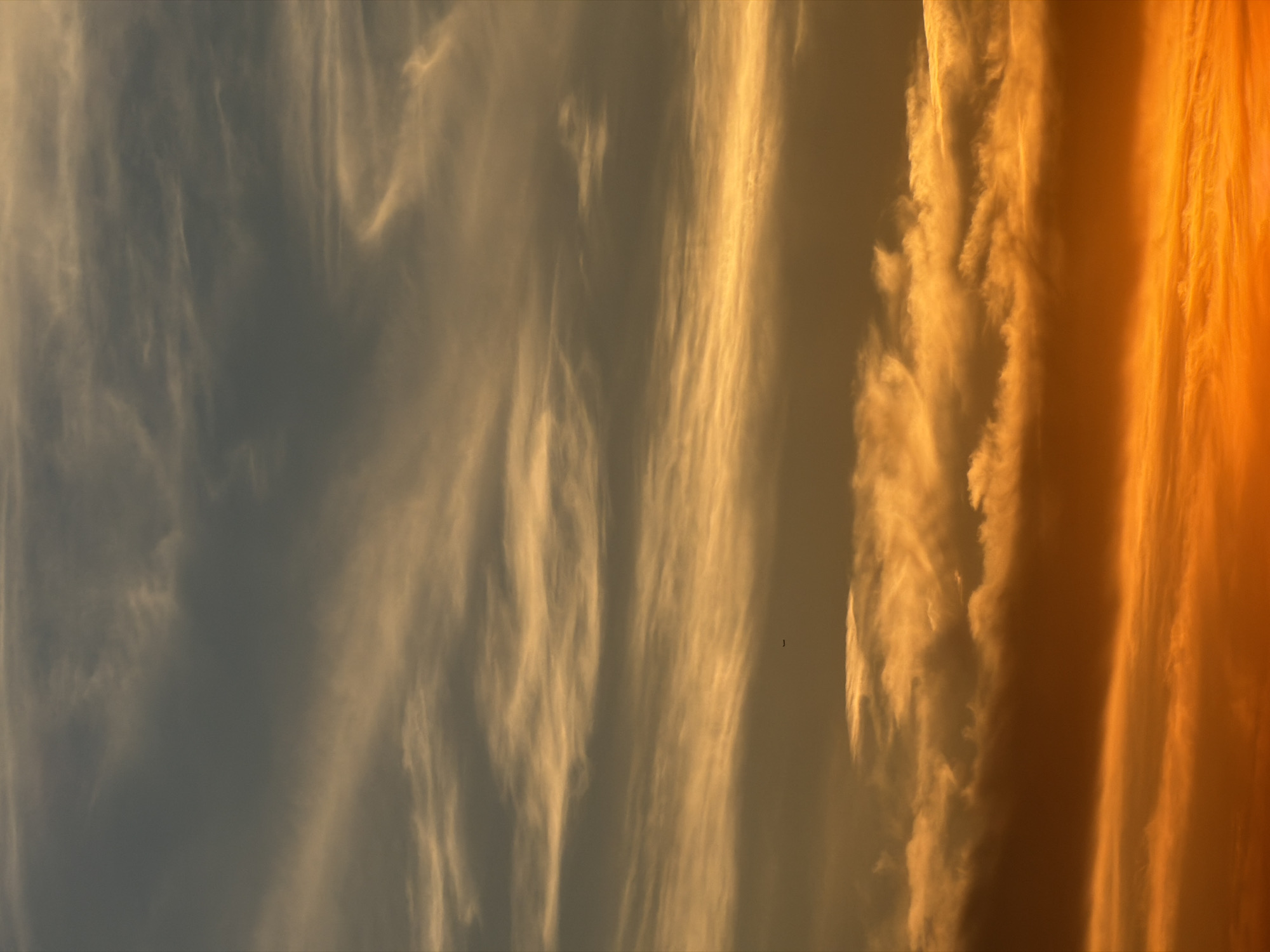 The image displays a vertical composition of a sky dominated by cloud formations, indicative of either sunrise or sunset, in the city of Unknown, Unknown. The scene features a distinct vertical color gradient.
The uppermost section of the frame consists of a dark greyish-blue sky containing thin, wispy, and somewhat translucent clouds that exhibit subtle golden illumination. These clouds appear streaky and attenuated.
Below this, the sky transitions into a prominent band of golden-yellow light. Within this middle section, more defined, elongated, and stratified clouds are present, intensely illuminated from below. A small, dark, horizontally oriented silhouette, identified as an airplane, is visible in the upper-middle left quadrant of this golden band.
The lower portion of the image is characterized by increasingly dense and volumetric cloud structures. These clouds are brightly illuminated with vibrant orange hues on their undersides, casting subtle shadows on their upper surfaces. The lowest section of the frame is dominated by a rich, deep reddish-orange color, suggesting the presence of the sun just below the visible horizon. No human figures, structures, or ground features are present within the frame.