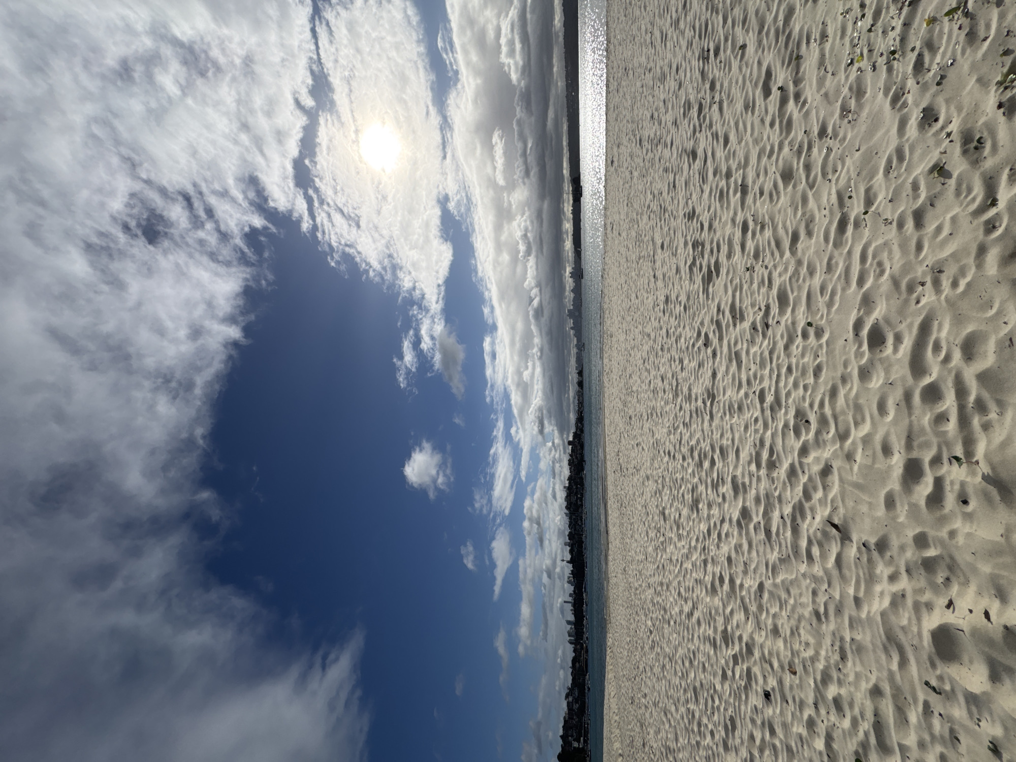 A wide, sandy beach stretches across the foreground of the image, its surface rippled by the wind. The sand is a pale, light beige color. In the mid-ground, a calm body of water, likely a bay or harbor, is visible. The water reflects the sunlight, creating a shimmering effect. Across the water, a city skyline is silhouetted against the horizon. Buildings of varying heights are visible, with some taller structures suggesting a downtown area. A bridge is also discernible on the far right of the skyline. Several boats are scattered across the water, some appearing to be sailboats. The sky above is a vibrant blue, punctuated by scattered white clouds. The sun is visible, partially obscured by clouds, casting a bright light. The scene appears to be set in a coastal urban environment, consistent with the city of Vaucluse, Australia, known for its harbor views and beaches.