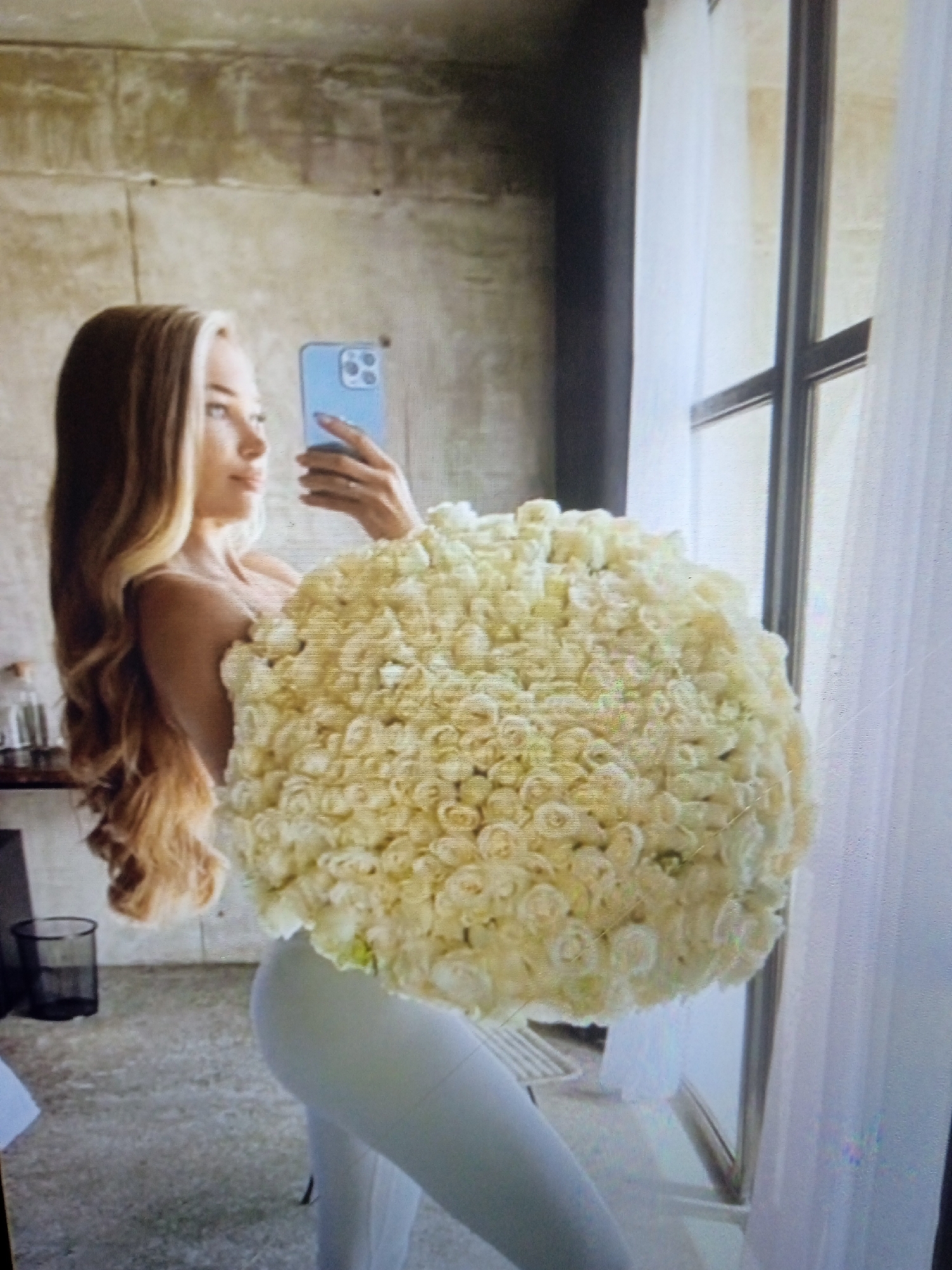 A person is taking a selfie with a large bouquet of white roses. The person is holding a blue smartphone up to take the photo. The person has long blonde hair and is wearing a white top and white pants. The background appears to be an indoor setting with a concrete wall and a large window with white curtains. There is a trash can on the left side of the image. The scene does not provide any specific details that would indicate a location in Chernivtsi, Ukraine.