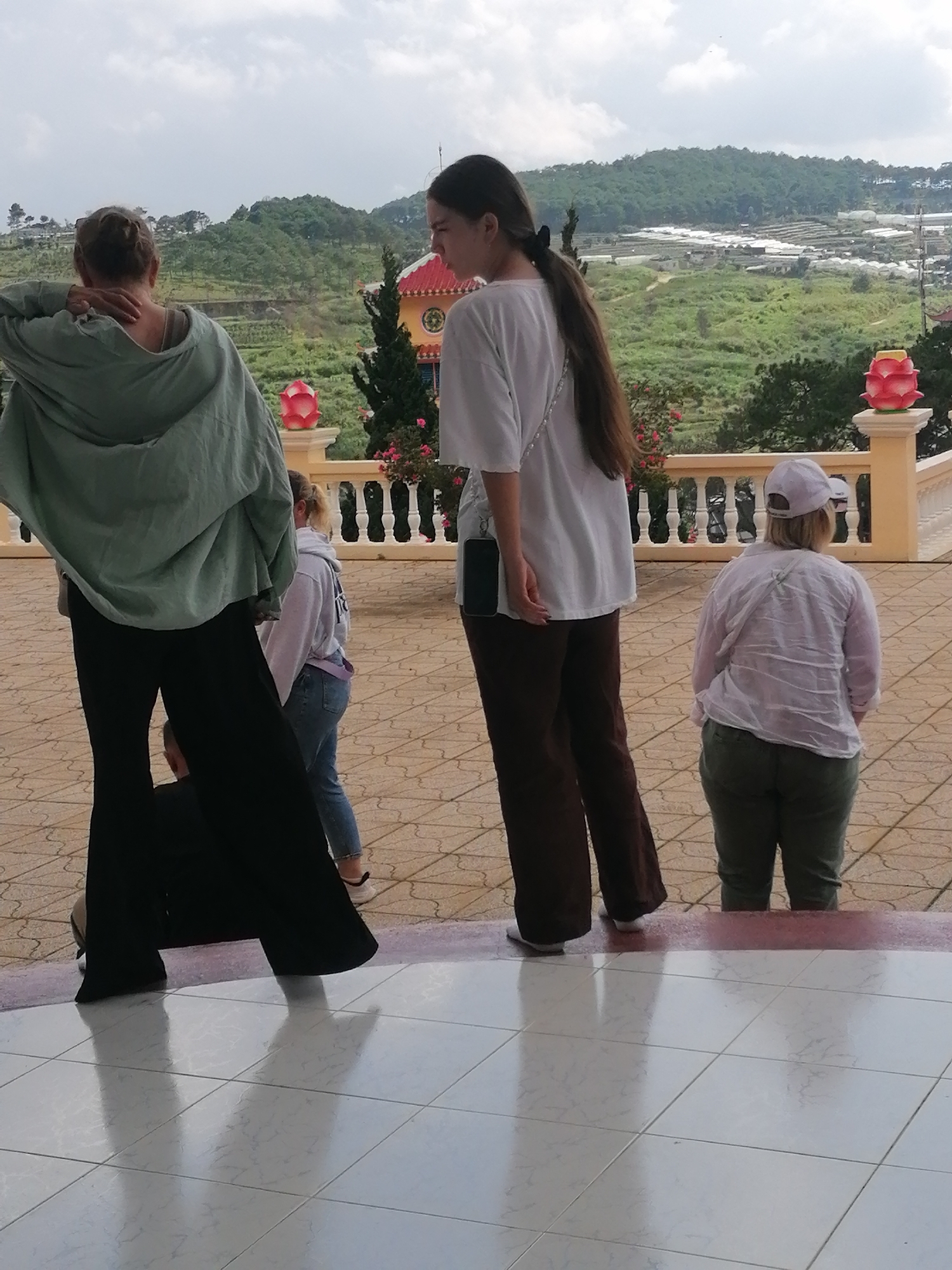 The image captures an outdoor scene with a view of rolling hills and agricultural terraces, likely in Lộc Quý, Vietnam. The foreground features a tiled walkway and steps. Four individuals are visible. On the left, a person wearing a light green hooded jacket and black flared pants stands with their back to the camera, reaching up to their neck. To their right and slightly behind, another person in a light purple hoodie and blue jeans is also seen from the back, facing away from the camera. In the center, a person in a white t-shirt and dark brown pants stands facing away, with a phone attached to their waistband. Further to the right, a person wearing a white long-sleeved shirt and green pants, with a white baseball cap, is also seen from the back. A decorative railing with vertical balusters and ornate pillars topped with pink lotus-like ornaments borders the mid-ground. The background reveals a vast landscape of green hills, patterned with what appear to be agricultural plots and greenhouses, under a cloudy sky.