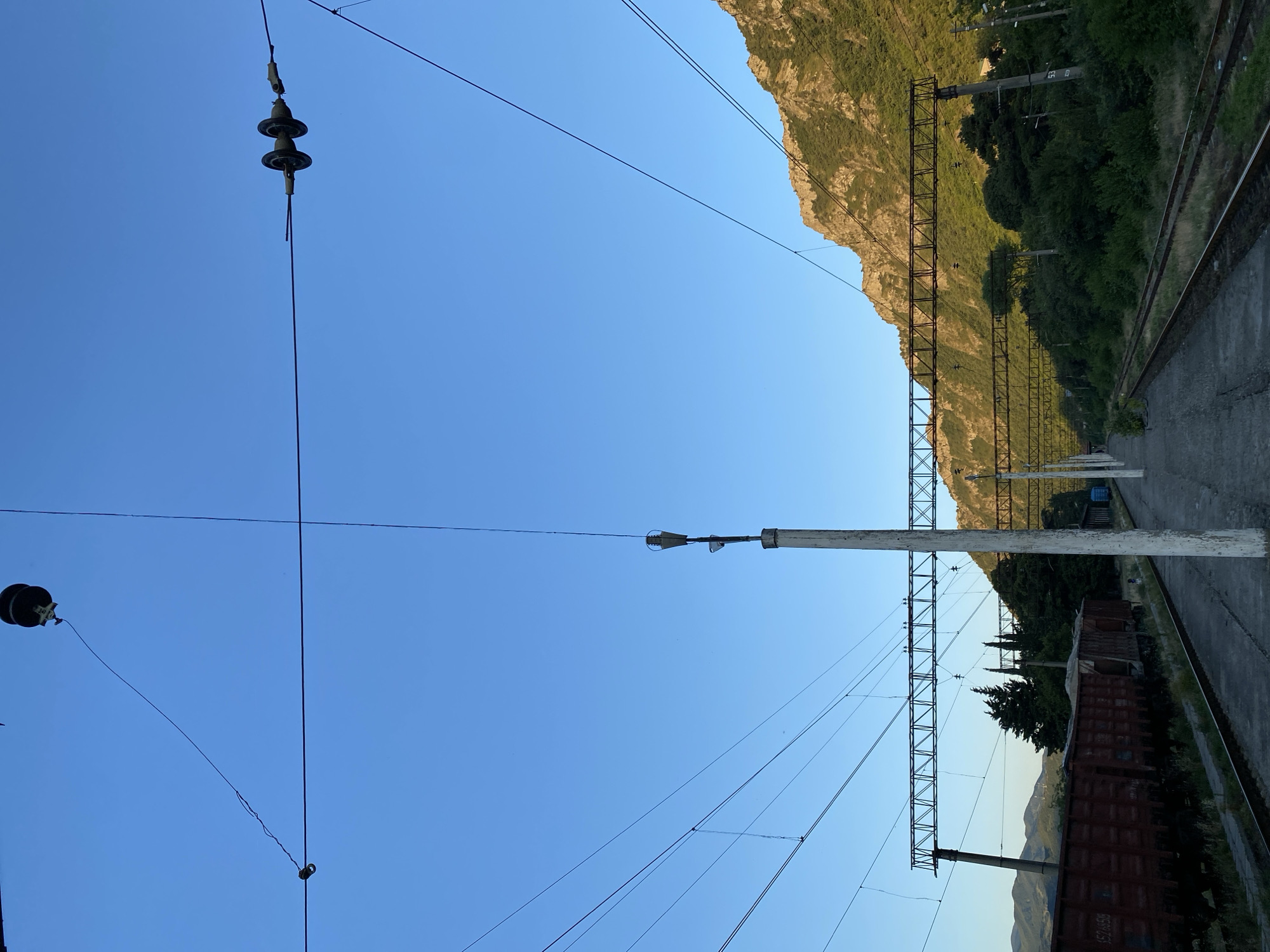 This image depicts a railway station in Mtskheta, Georgia. The scene is dominated by the clear blue sky and overhead power lines for an electric train. A rugged, tree-covered mountain range rises in the background, catching the warm light of what appears to be late afternoon or early evening.
In the foreground, on the right side of the image, are railway tracks and a concrete platform. To the left of the tracks, a rusty freight train car is parked. The train car has some text visible, but it's difficult to read clearly in the image. Along the track, there are also utility poles supporting the overhead wires, typical of an electrified railway system. One pole has the number "53" printed on it.
The overall impression is one of a tranquil, somewhat remote railway station nestled in a mountainous landscape. The lighting suggests a peaceful and calm atmosphere. There are no people visible in the frame.
