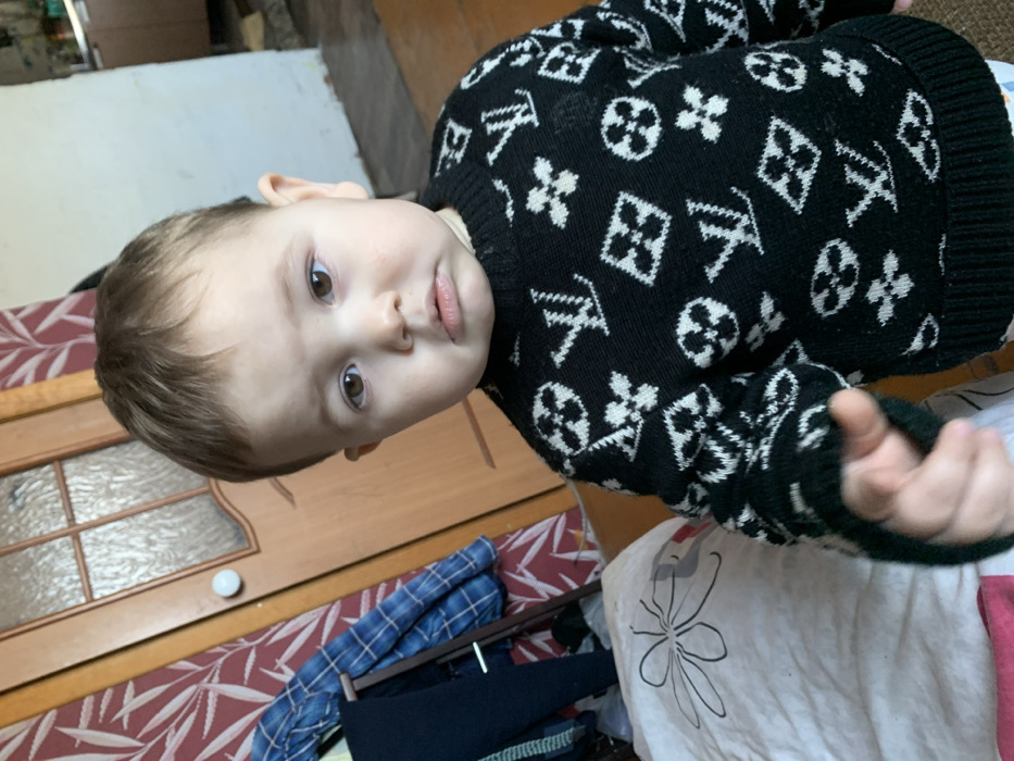 An indoor image captured in a residence in Фоки, Russia, features a toddler-aged child positioned centrally. The child has light brown hair, brown eyes, and a serious, slightly pouting facial expression. They are dressed in a black knitted sweater adorned with repeating white patterns resembling the Louis Vuitton monogram, featuring a dark green ribbed hem. A white and light blue striped garment is visible beneath the sweater. The child's right hand is slightly extended towards the camera, with fingers loosely curled.
The background on the left side of the frame includes a wall covered in reddish-brown wallpaper with lighter botanical patterns, a partially visible light-brown wooden door with frosted glass panels and a white spherical doorknob. A clothes rack holding blue plaid and dark-colored garments stands to the left of the door. In the lower left foreground, a white fabric item with a black outline of a floral design is present. The background on the right reveals a plain light-colored wall and a light-brown wooden floor. Faint light-colored kitchen cabinetry is visible in the distant background on the right. The scene is illuminated by ambient indoor light.