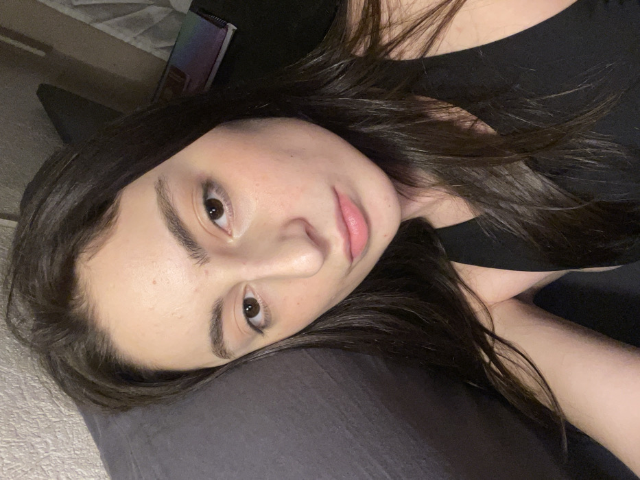 The scene is situated indoors, within what appears to be a residential environment in Moscow, Russia. The image features a single female subject, positioned reclining or lying down, with her head resting on a dark gray pillow or cushion. She has long, dark brown hair, which extends over her right shoulder and arm. Her complexion is fair, with visible pores and subtle redness on the cheeks. Her eyes are brown, and her eyebrows are dark and prominent. Her lips are naturally pink and slightly parted. She is clad in a black tank top. The subject's gaze is directed towards the camera, her expression neutral. In the background, a light-colored, textured wall is visible to the upper left. To the right of the subject's head, a dark surface is present, upon which a multi-colored, spiral-bound notebook or similar object is partially visible. The lighting is artificial, casting a soft illumination on the subject's face.
