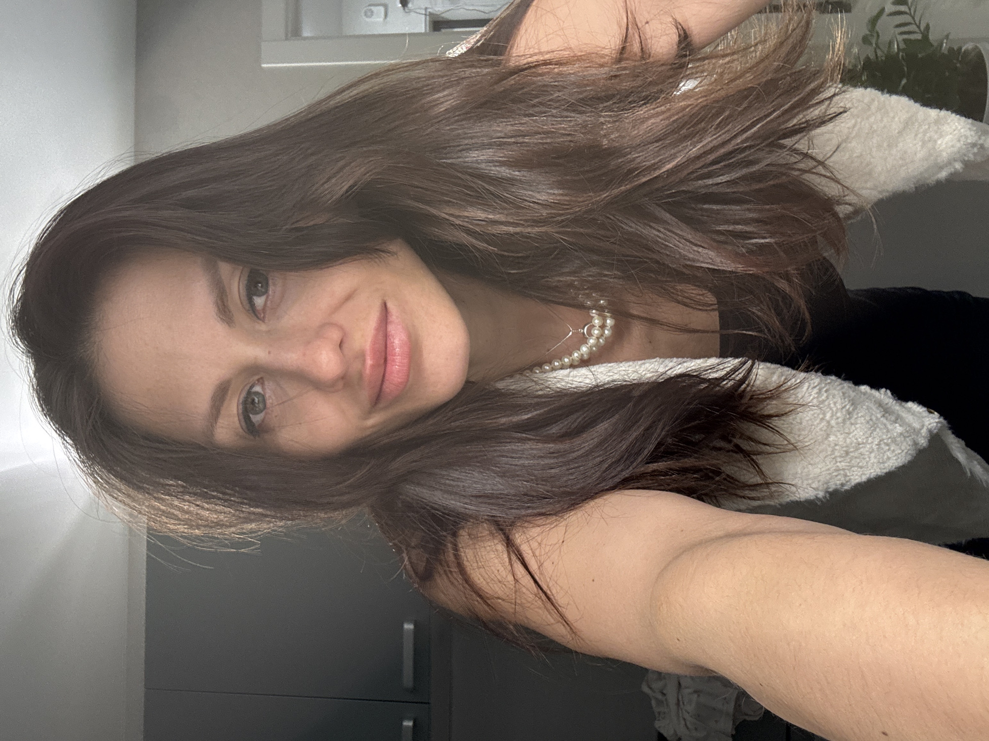 A selfie shows a woman with long, dark brown hair, light skin, and green eyes. She is smiling gently at the camera. She appears to be indoors with grey cabinets in the background. She is wearing a black top and a white, fluffy vest. A pearl necklace is visible around her neck. There are plants in the background.