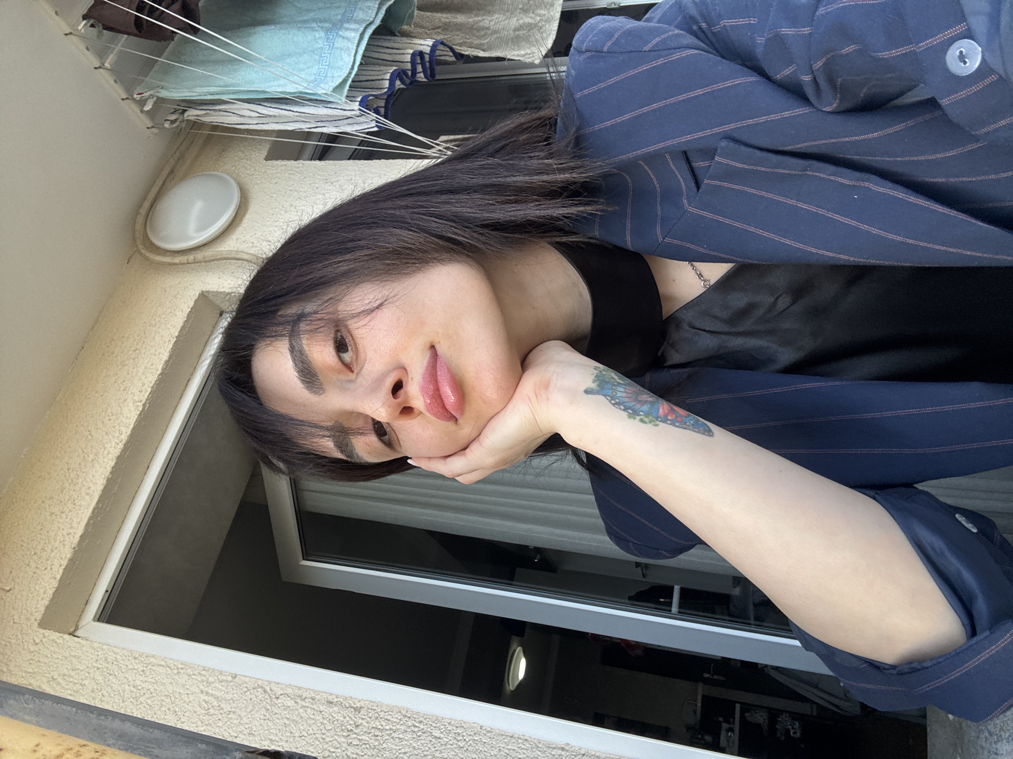 A person with long dark hair, wearing a navy pinstripe blazer and a black top, is resting their head on their hand. The person has a butterfly tattoo on their forearm. They are positioned in front of a window, with clothes hanging on a line visible in the background. The location is likely a residential building in Tbilisi, Georgia, based on the architectural style and the presence of a small balcony.