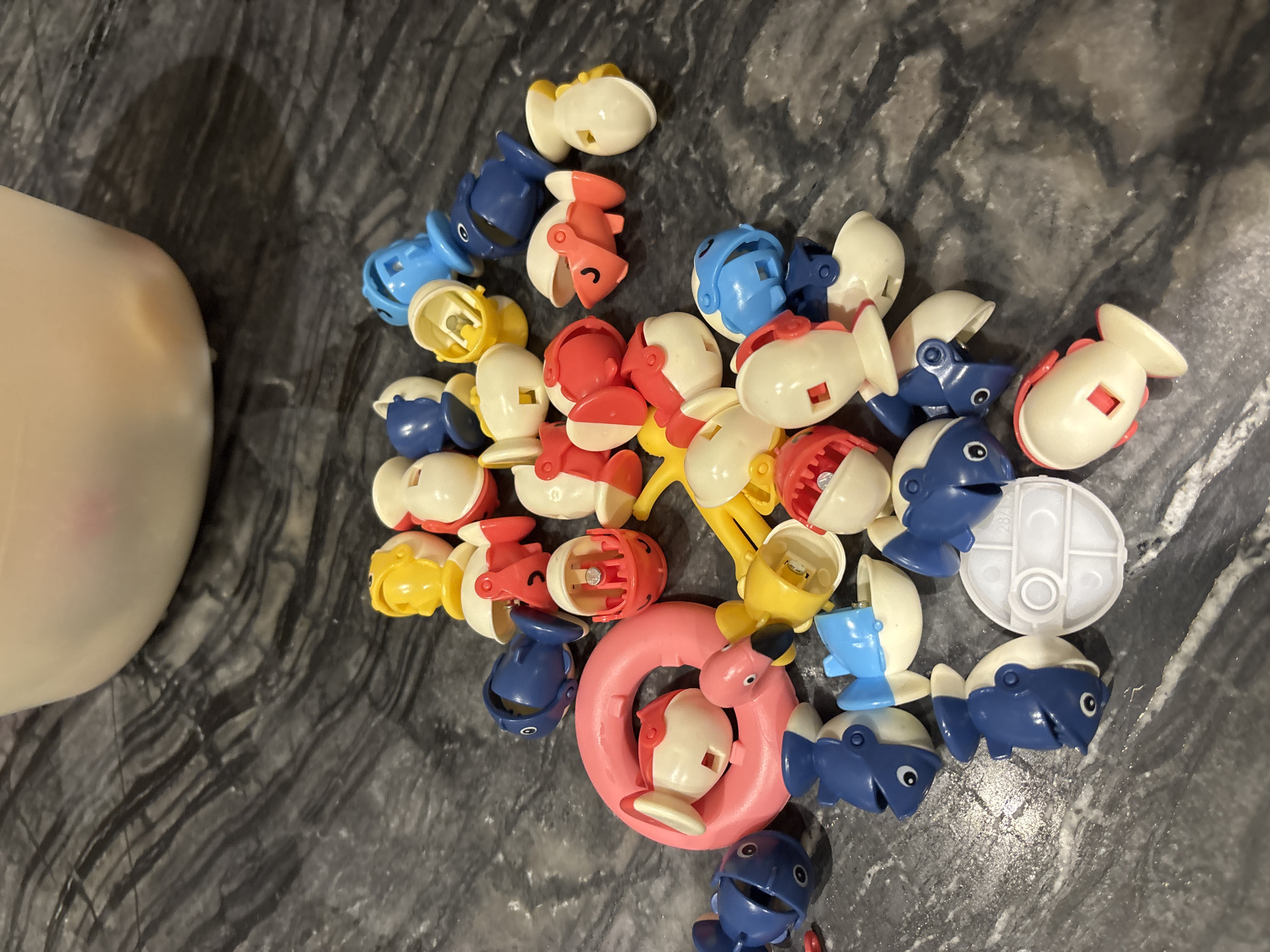 The image shows a collection of small, colorful plastic toys, primarily fish and egg-shaped figures, scattered on a dark, marbled surface. The toys are in various states of assembly, with some pieces detached from others. Dominant colors include blue, red, yellow, white, and pink. A large, out-of-focus white bowl or container is visible in the upper portion of the image. There are no people or signs of human presence, other than the scattered toys. The environment appears to be indoors, given the lighting and the surface material. There is no identifiable information in the image that would indicate its location in Dubai, United Arab Emirates.
